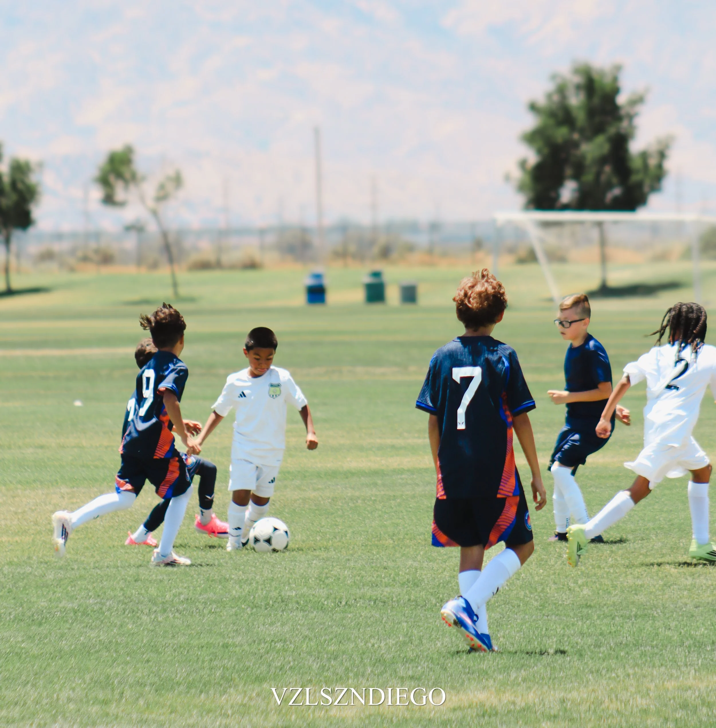 Youth soccer game on a sunny day with children in blue and white jerseys, children are running towards the soccer ball on the grass field. In the background, there are trees, a water tower, and a partly cloudy sky.