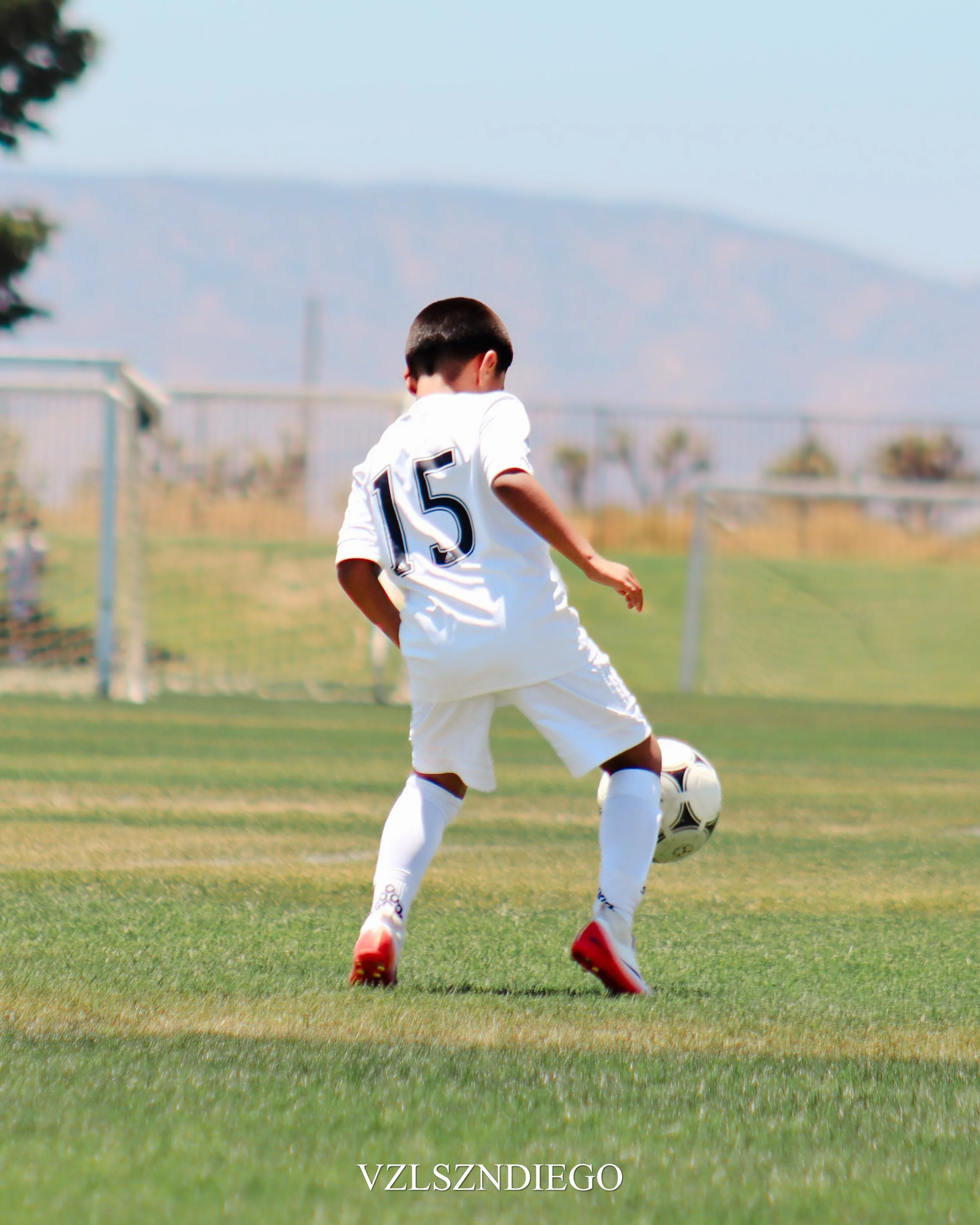 A boy wearing a white soccer uniform with the number 15 on the back, playing soccer on a field with a mountain in the background. The boy is preparing to kick or pass the ball, with one foot near the ball and the other foot planted on the ground.