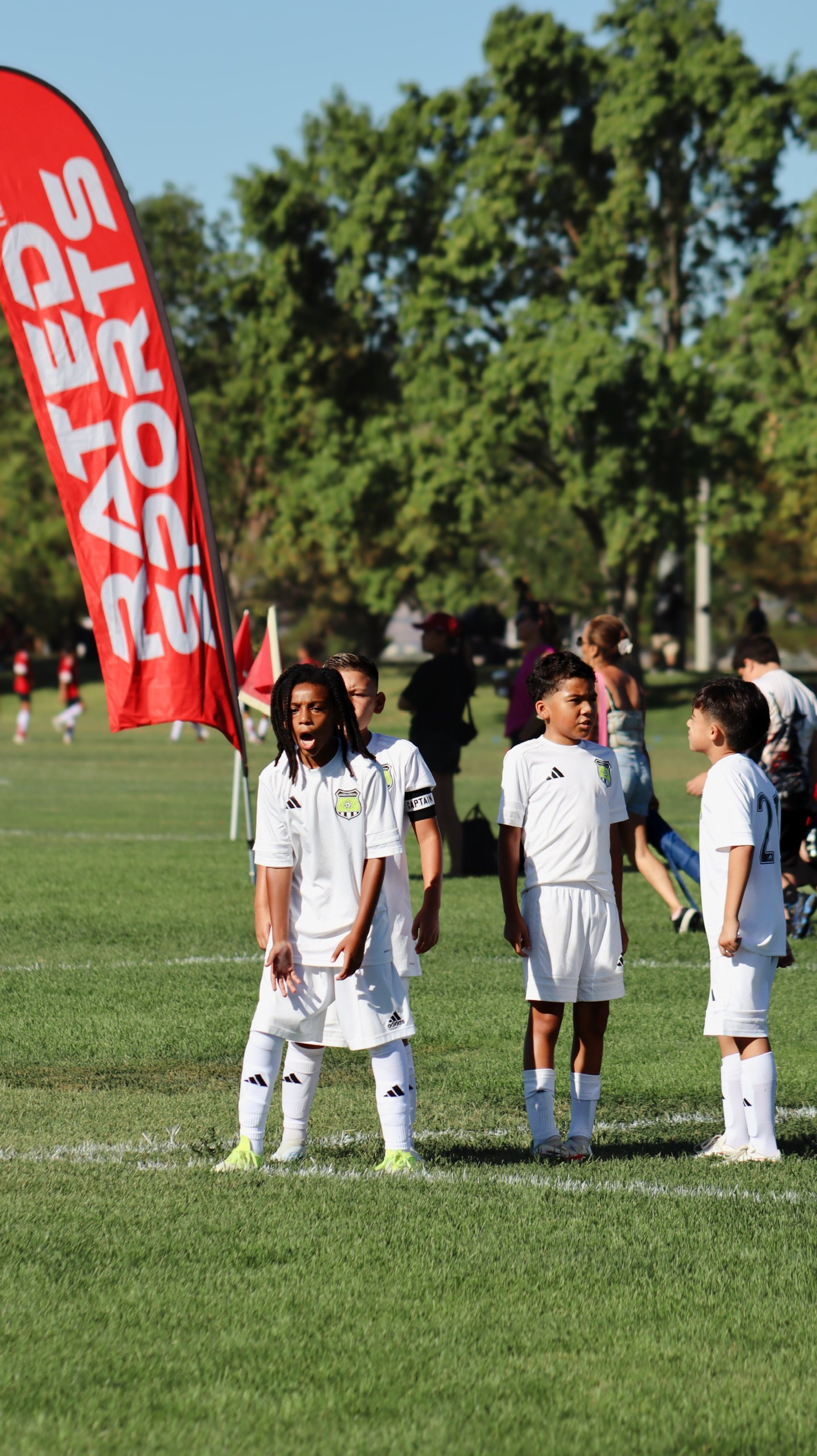Children in white soccer uniforms standing on a grassy field, with some people in the background, and a red flag with white text.
