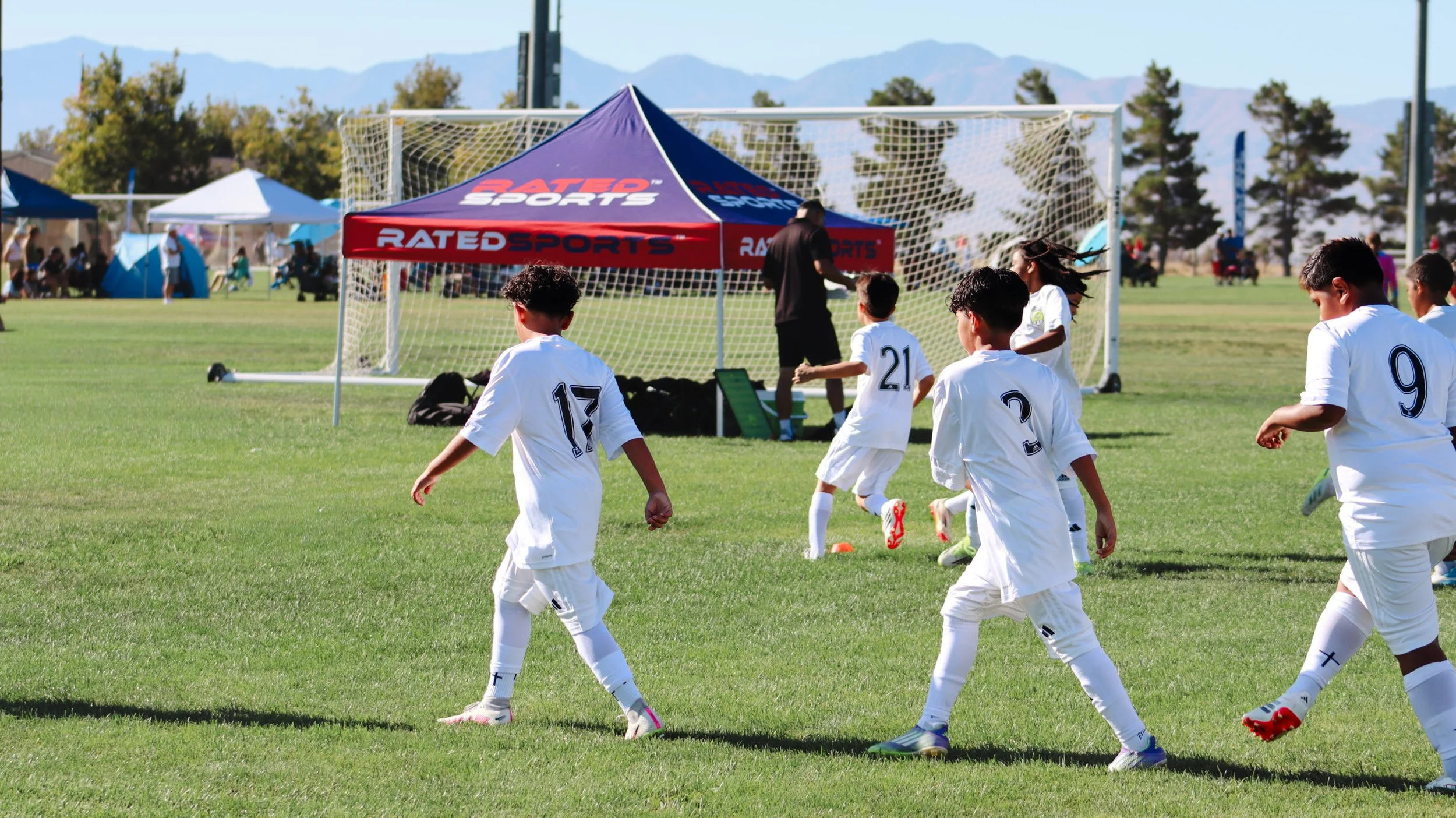 Young soccer players in white uniforms playing on a field during a game, with a tent marked 'RATED SPORTS' and a soccer goal in the background.