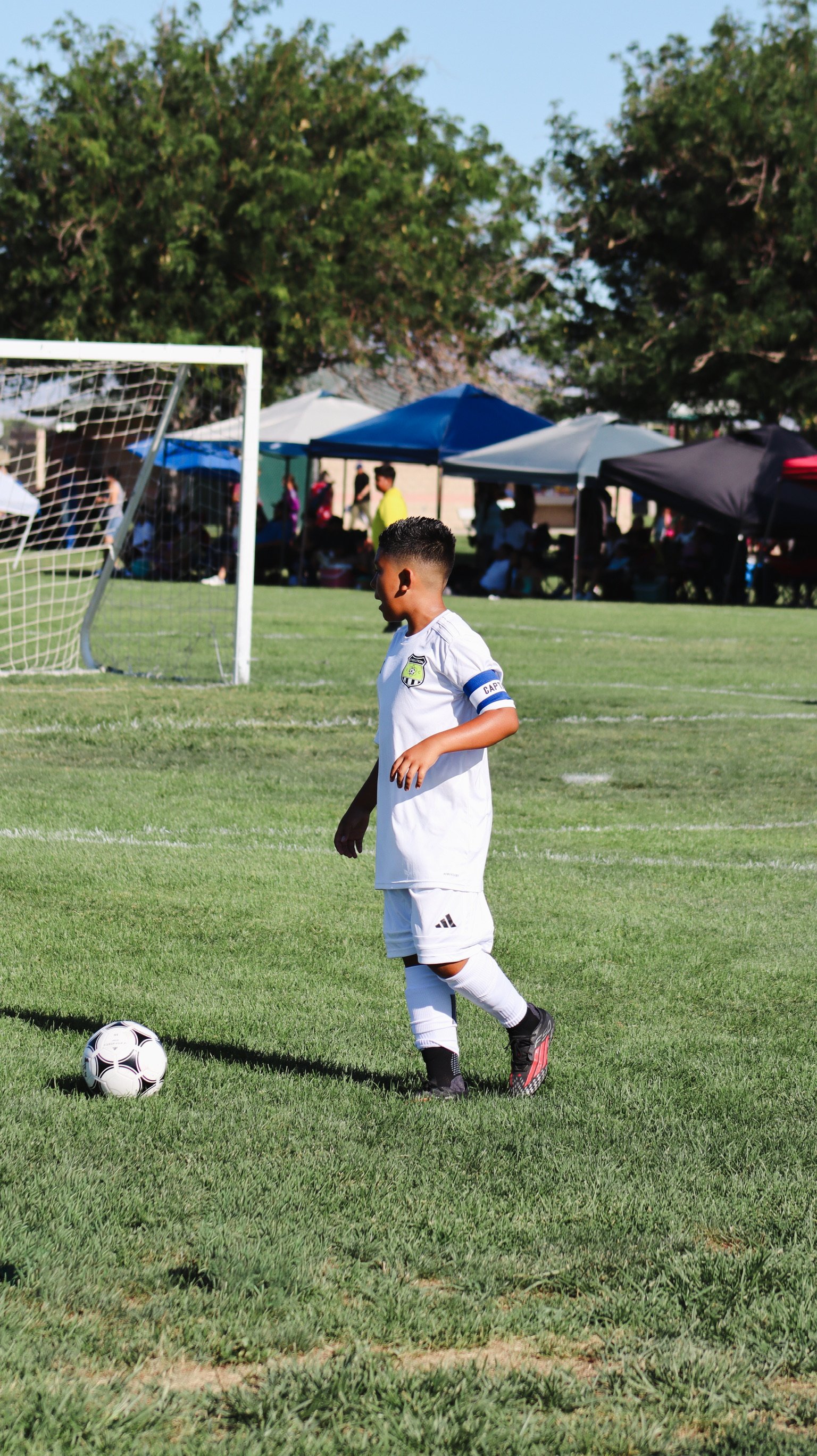 A young soccer player in a white uniform with a captain's armband and a logo on the chest, standing on a grassy field near a soccer ball, with tents and spectators under trees in the background on a sunny day.