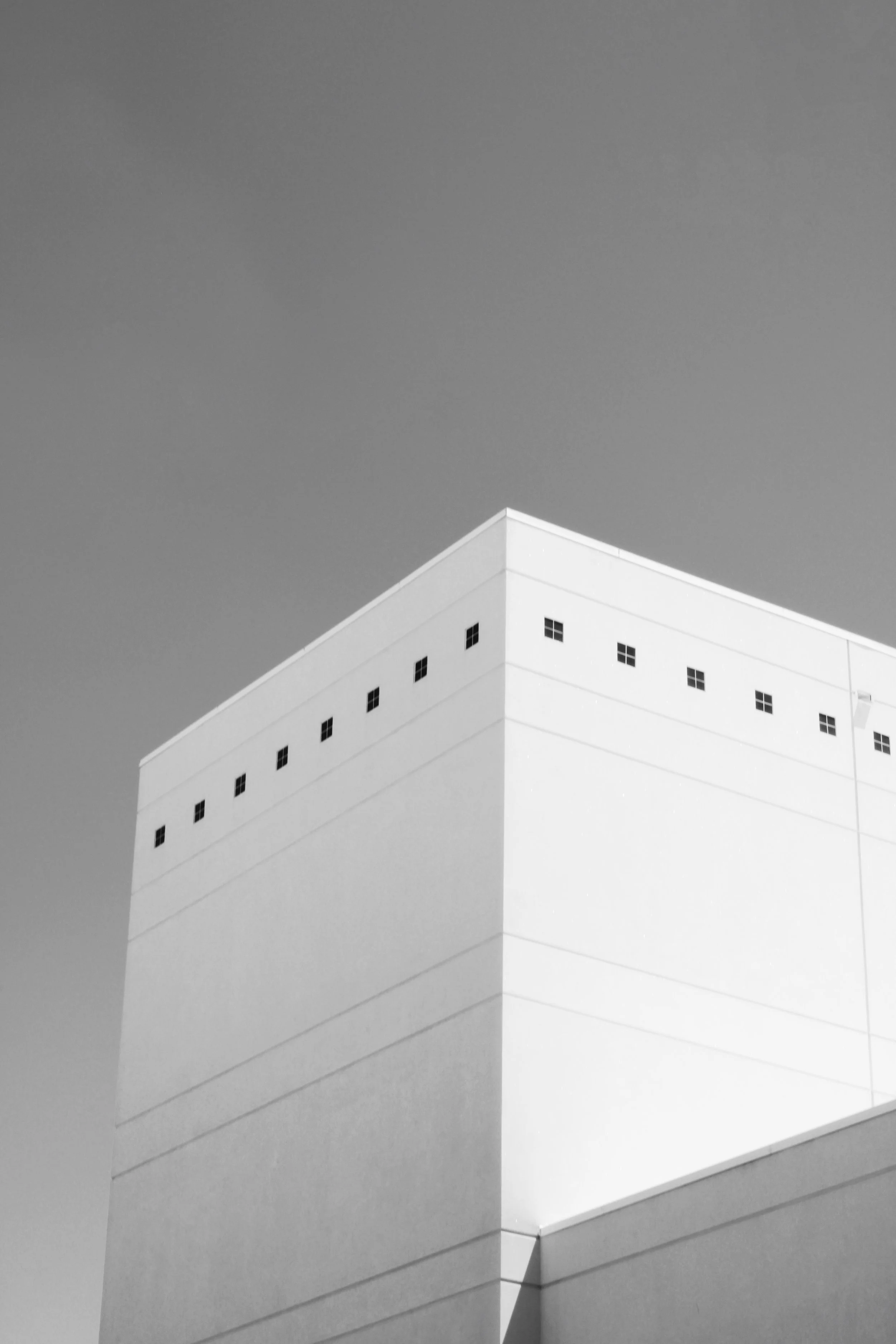 Close-up black and white photograph of the corner of a modern white building with small square windows, captured from a low angle against a clear sky.