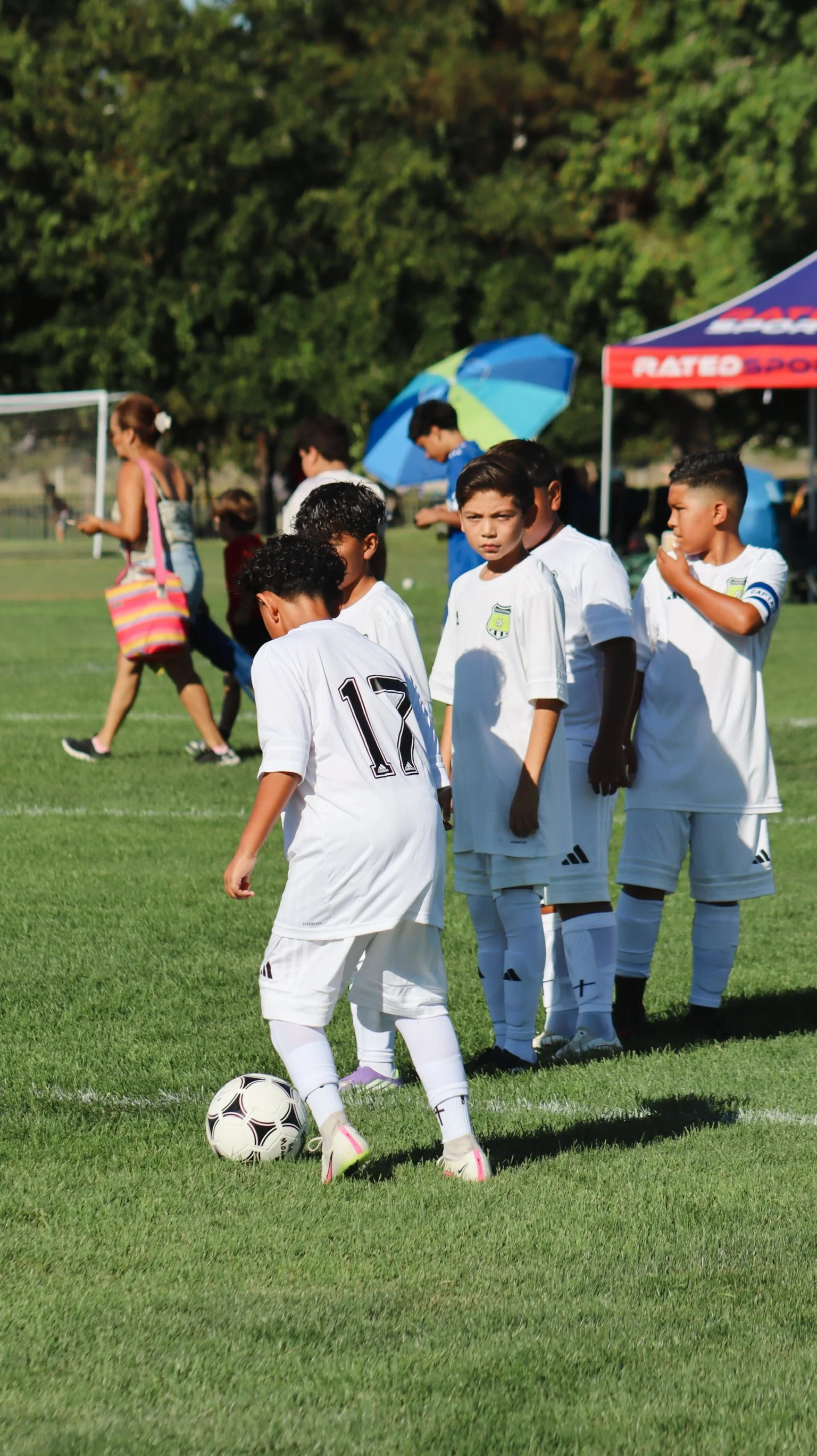 Children wearing white soccer uniforms are standing on a grassy field, with one child about to kick a soccer ball. There are adults and umbrellas in the background, with trees and a tent.