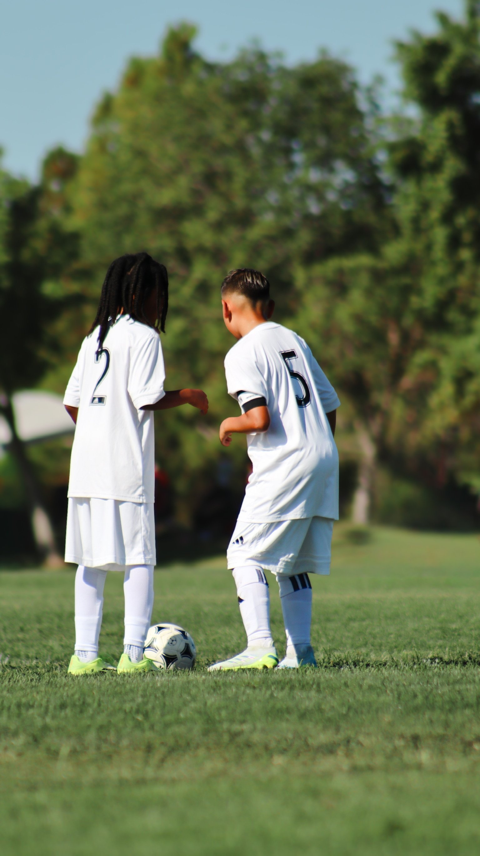 Two children in soccer uniforms standing on a grassy field with a soccer ball, one girl with dreadlocks and one boy with short hair, in a park with trees in the background.