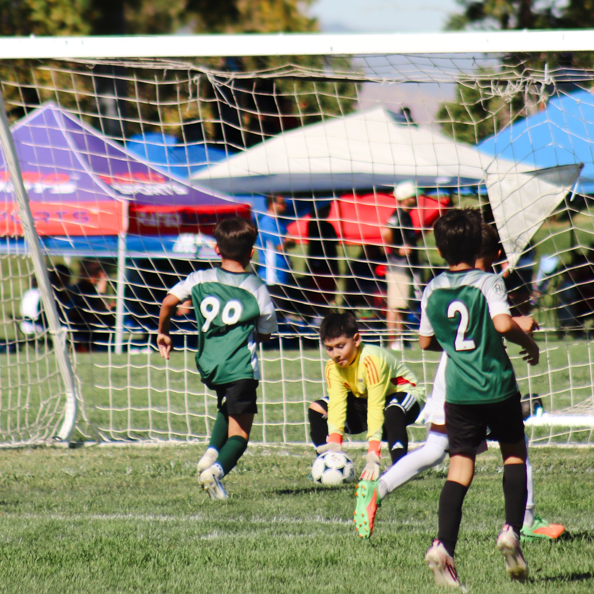 Children playing soccer on a grassy field, with a goalpost and net in the foreground, and tents and trees in the background during daytime.