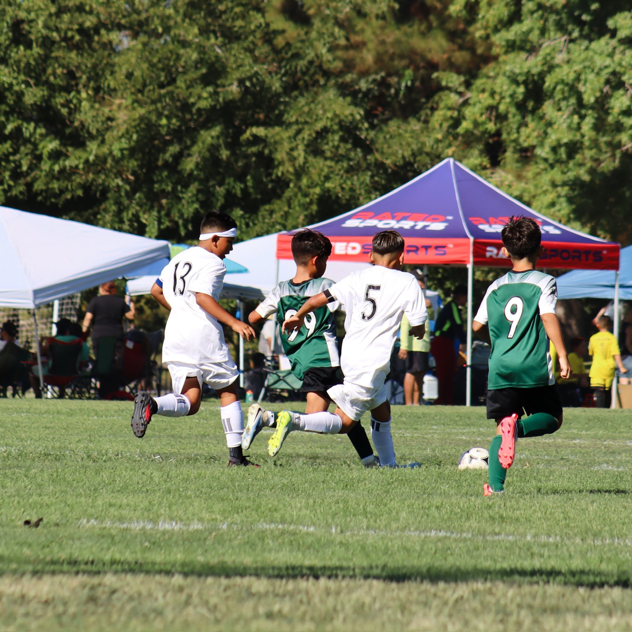 Soccer players in green and white uniforms on the field, with tents and trees in the background, participating in a match.