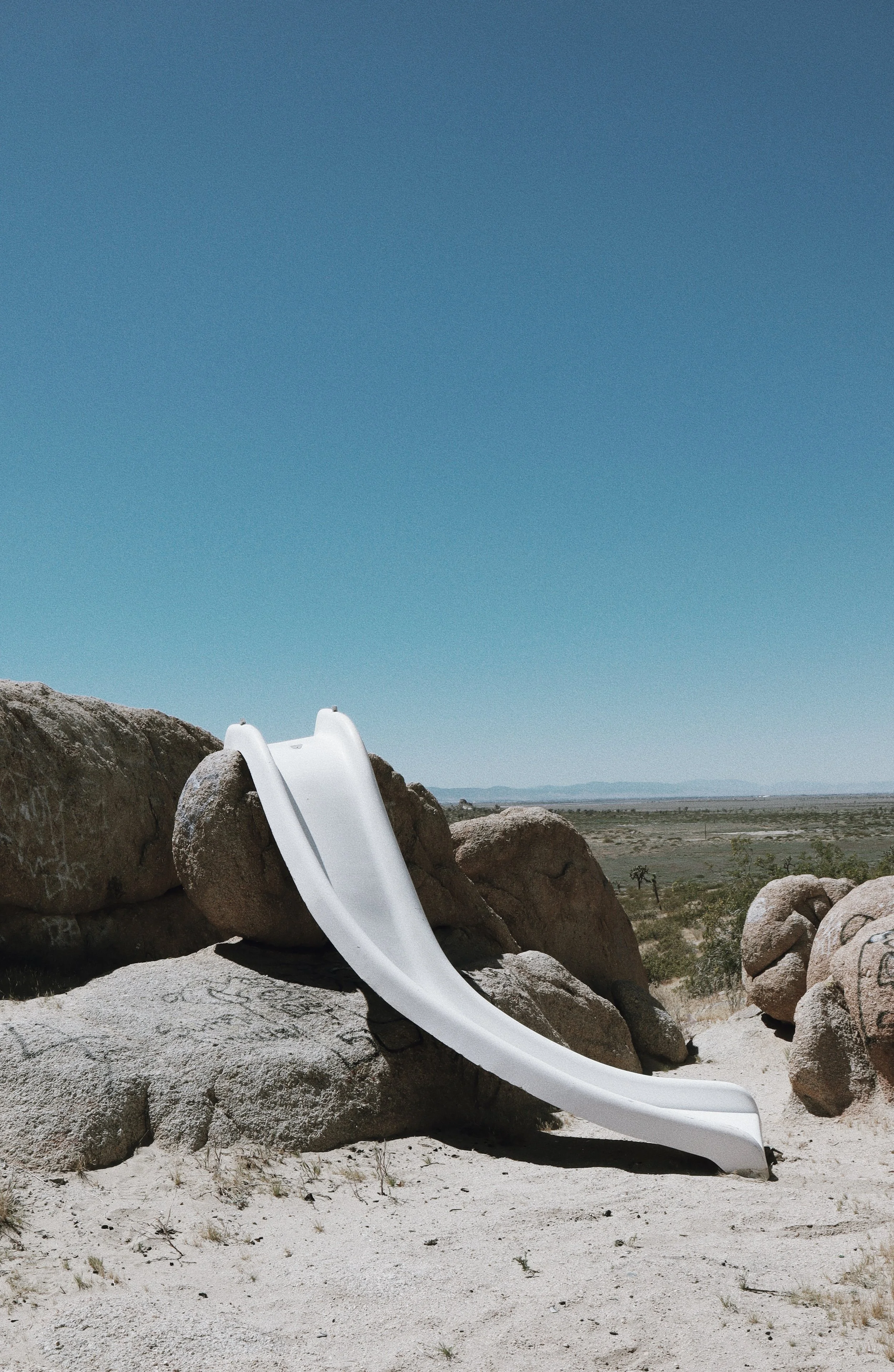 A small white water slide placed on rocks in a desert landscape with stones, sparse vegetation, and mountains in the background under a clear blue sky.