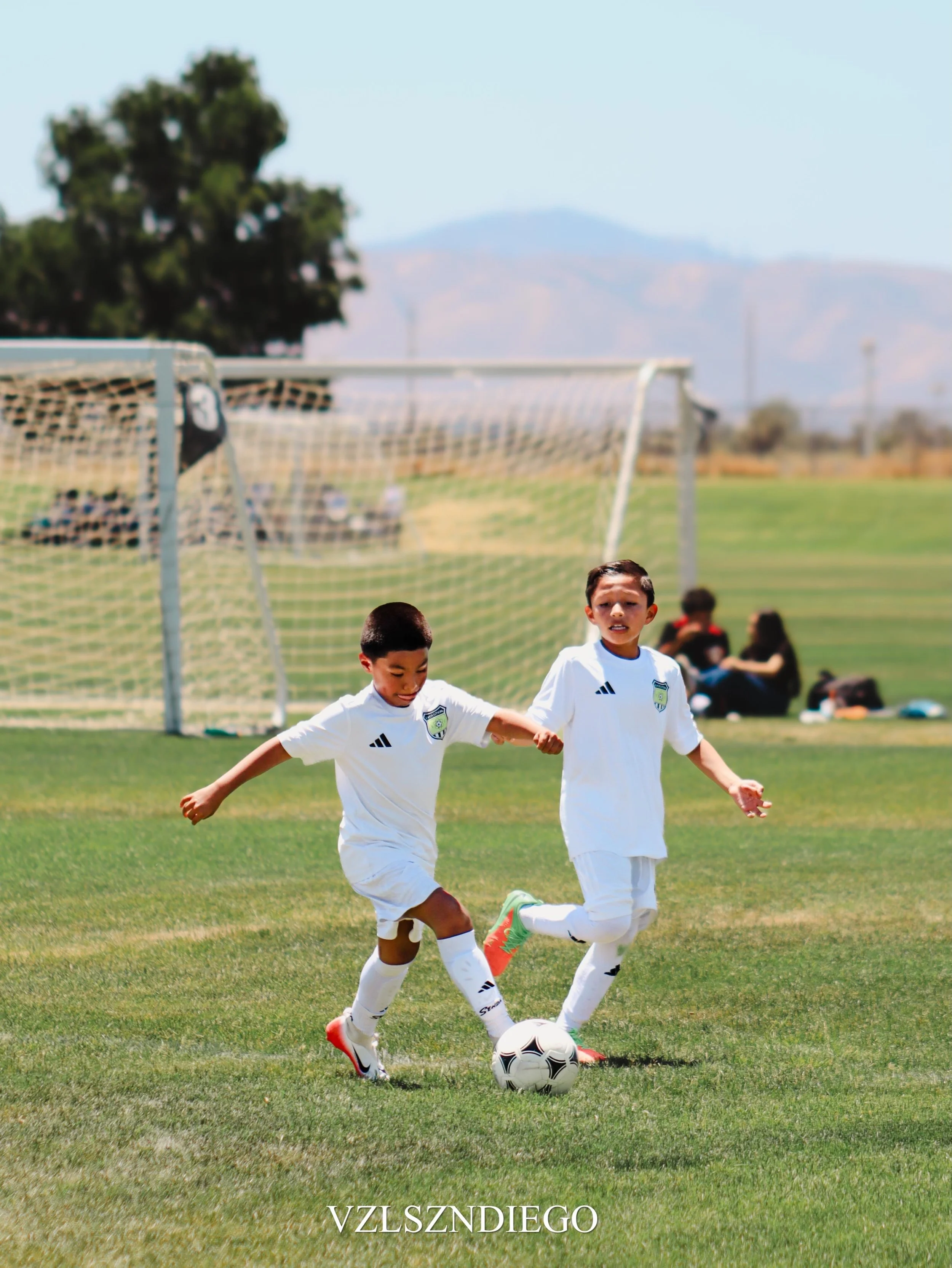 Two young boys wearing soccer uniforms are playing soccer on a green field. They are chasing a soccer ball with a fence, some people sitting on benches, a large tree, and mountains in the background. The word "VZLSZNDIEGO" is visible at the bottom of