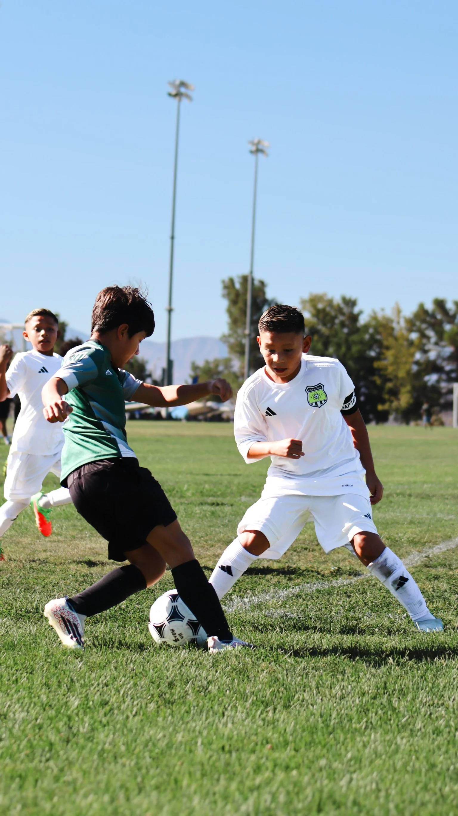 Young boys playing soccer on a sunny field, with some players focused on the ball and others running in the background, under a clear blue sky.