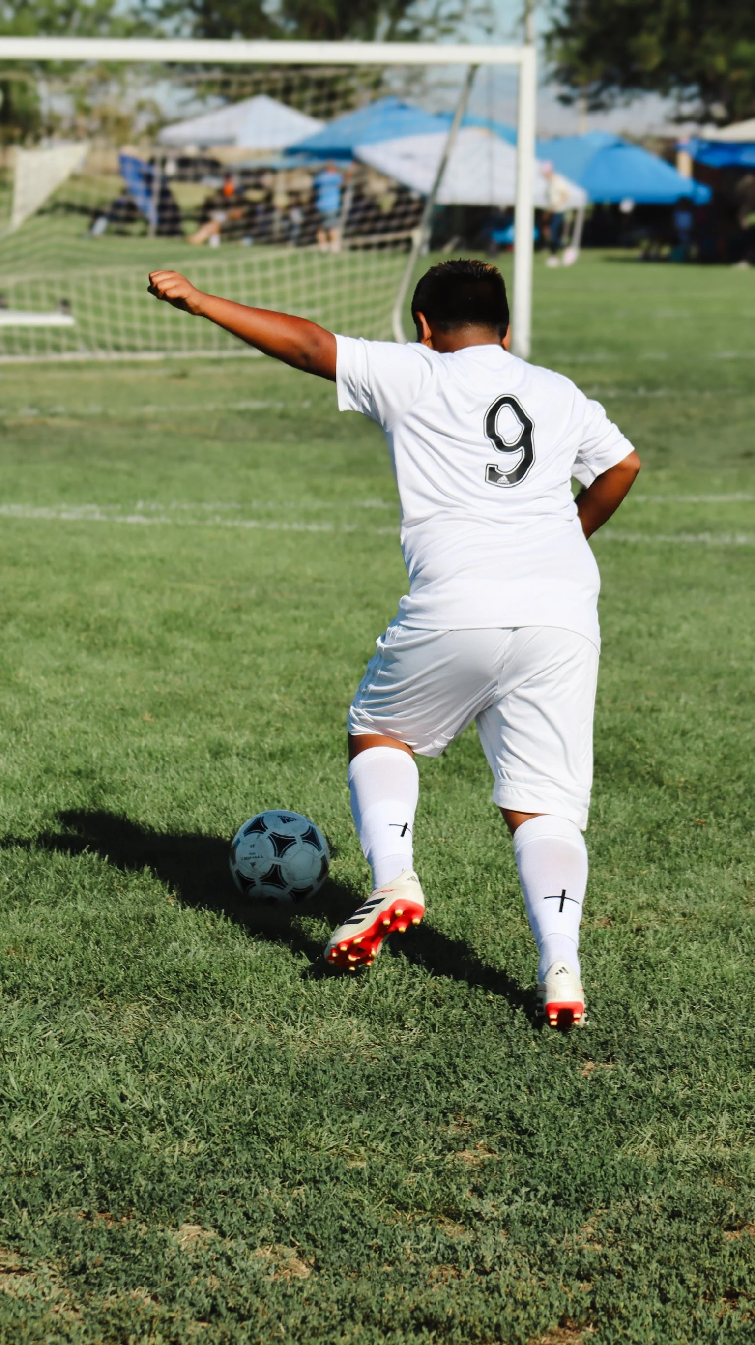 Soccer player wearing white jersey with number 9, kicking a soccer ball on a grassy field with goal post and spectators under tents in background.