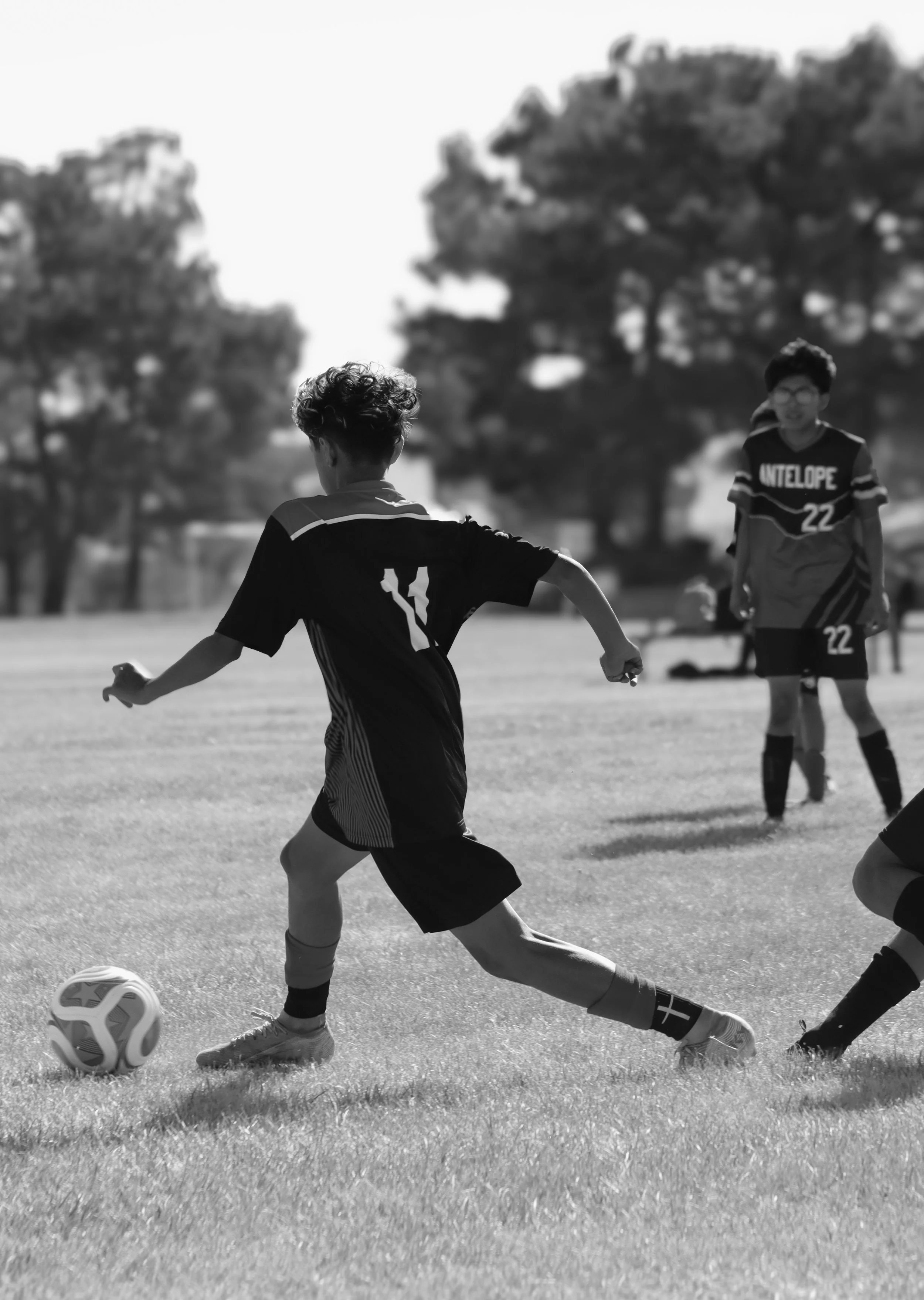 Two young boys playing soccer on a grassy field. One boy in the foreground is kicking a soccer ball, and the other boy in the background is smiling, wearing glasses and a jersey with the number 22.