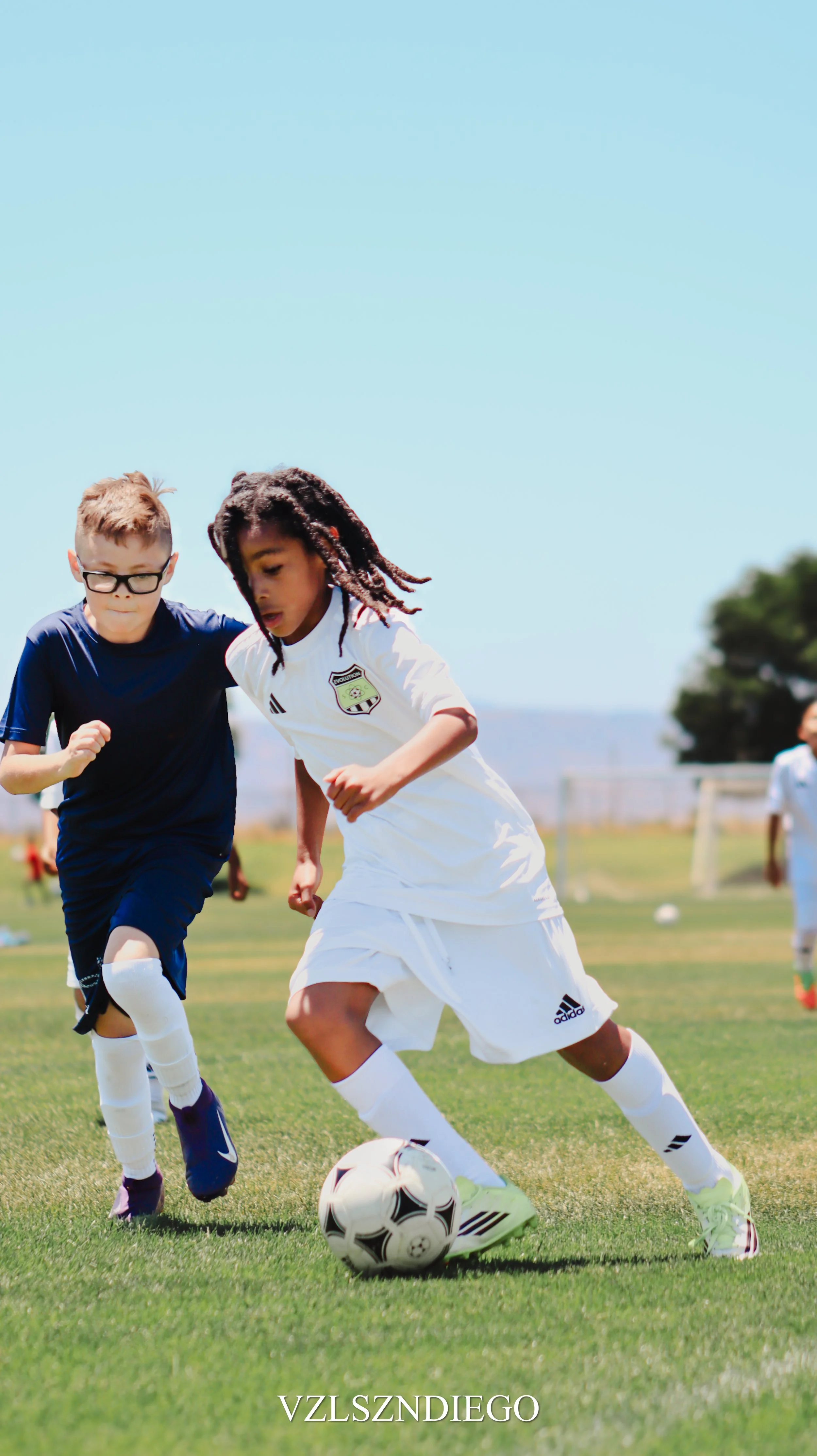 Two young boys playing soccer outdoors on a sunny day, both focused on the ball as they run on the grass field.