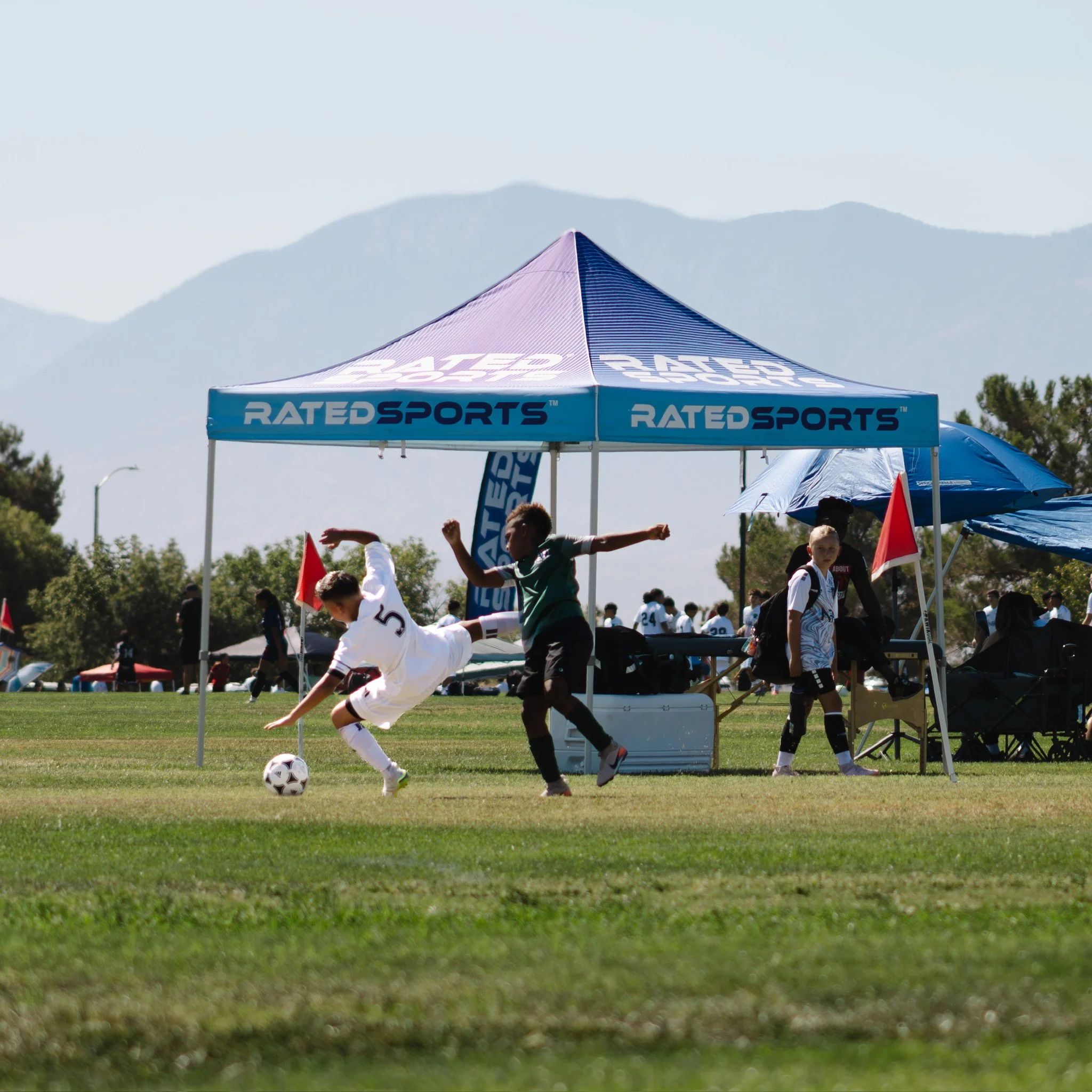 Children playing soccer under a blue sports tent in an outdoor field, with mountains in the background on a sunny day.