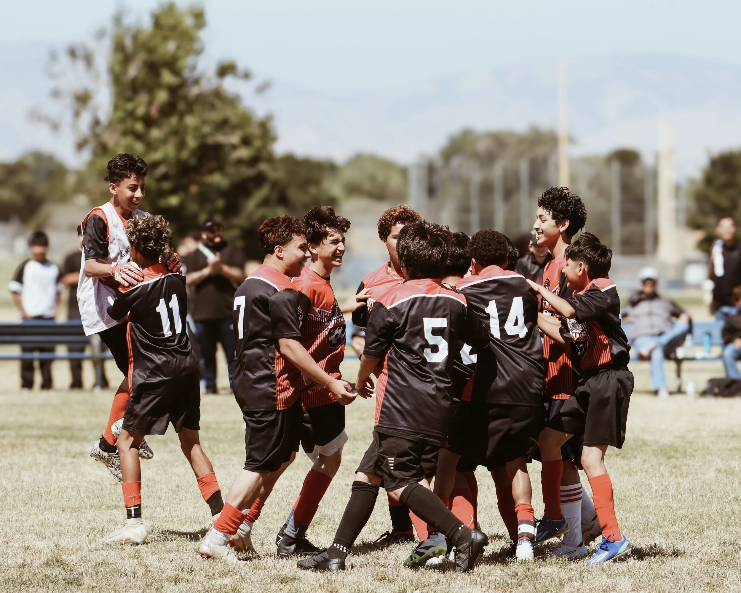 A group of young soccer players in black and red uniforms huddled together on a field, celebrating.