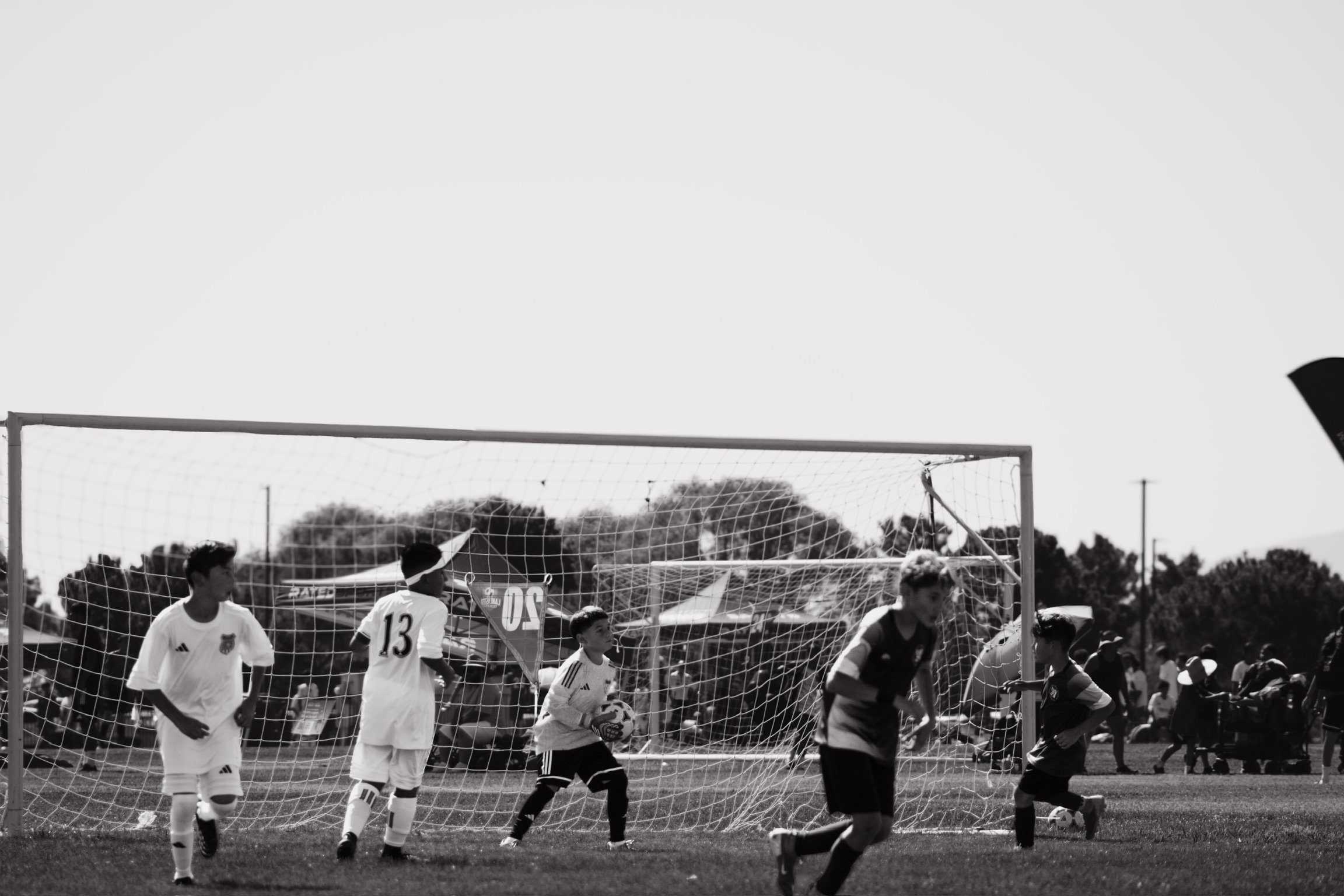 Black and white image of children playing soccer on a field, with a goalpost in the foreground and trees in the background.