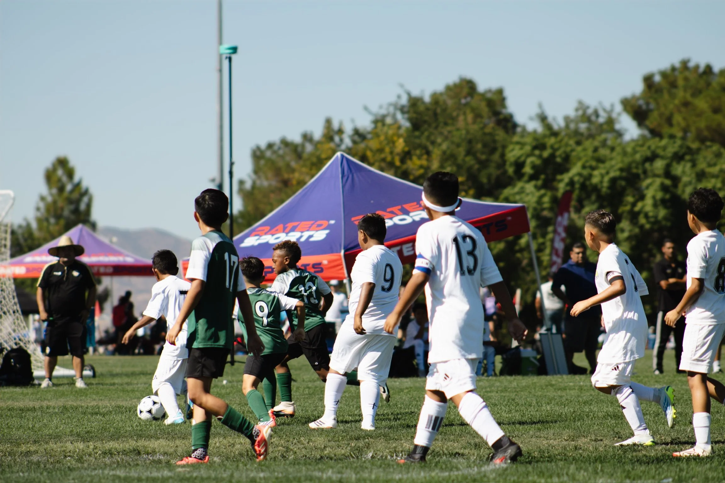 Young boys playing soccer at an outdoor field with family, coaches, and tents in the background.