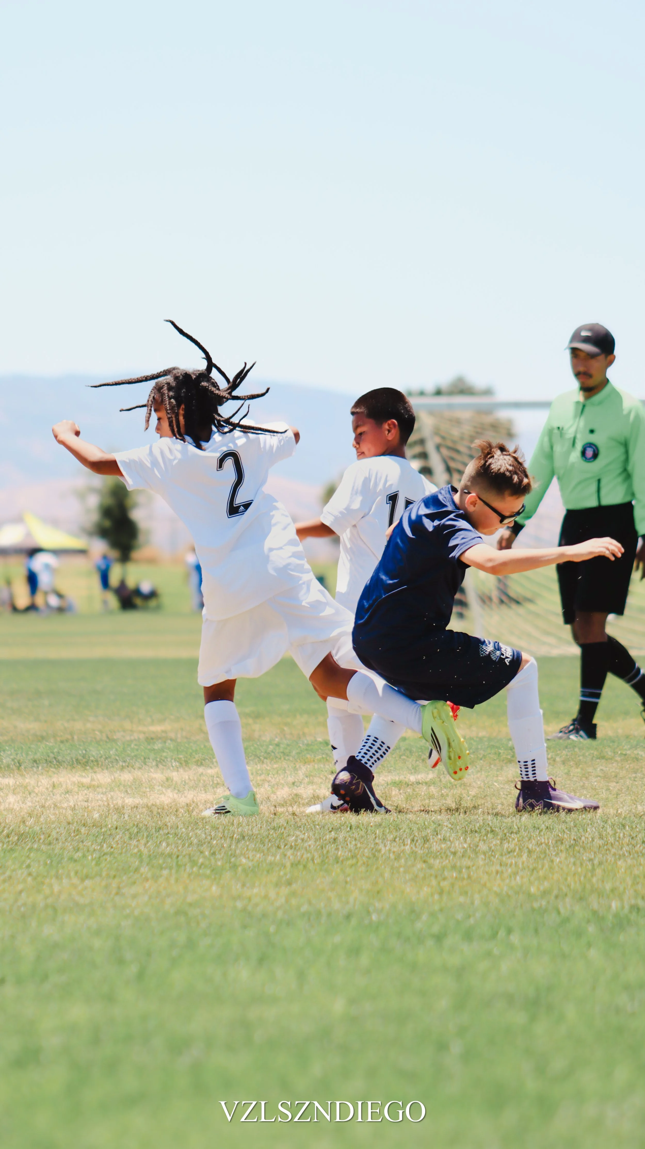 Children playing soccer on a grassy field with a referee in the background during daytime.