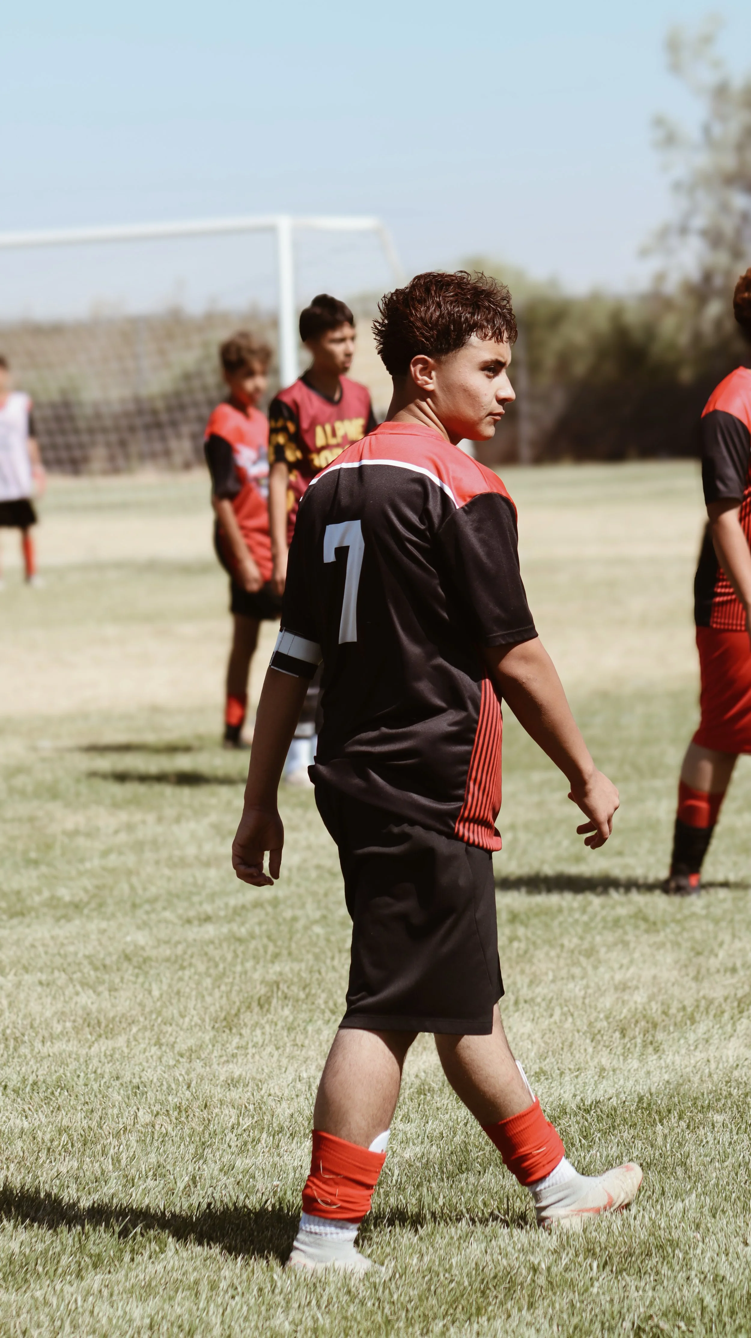 A young male soccer player with short curly hair, wearing a black and red uniform with the number 7, standing on a grassy field during a game.