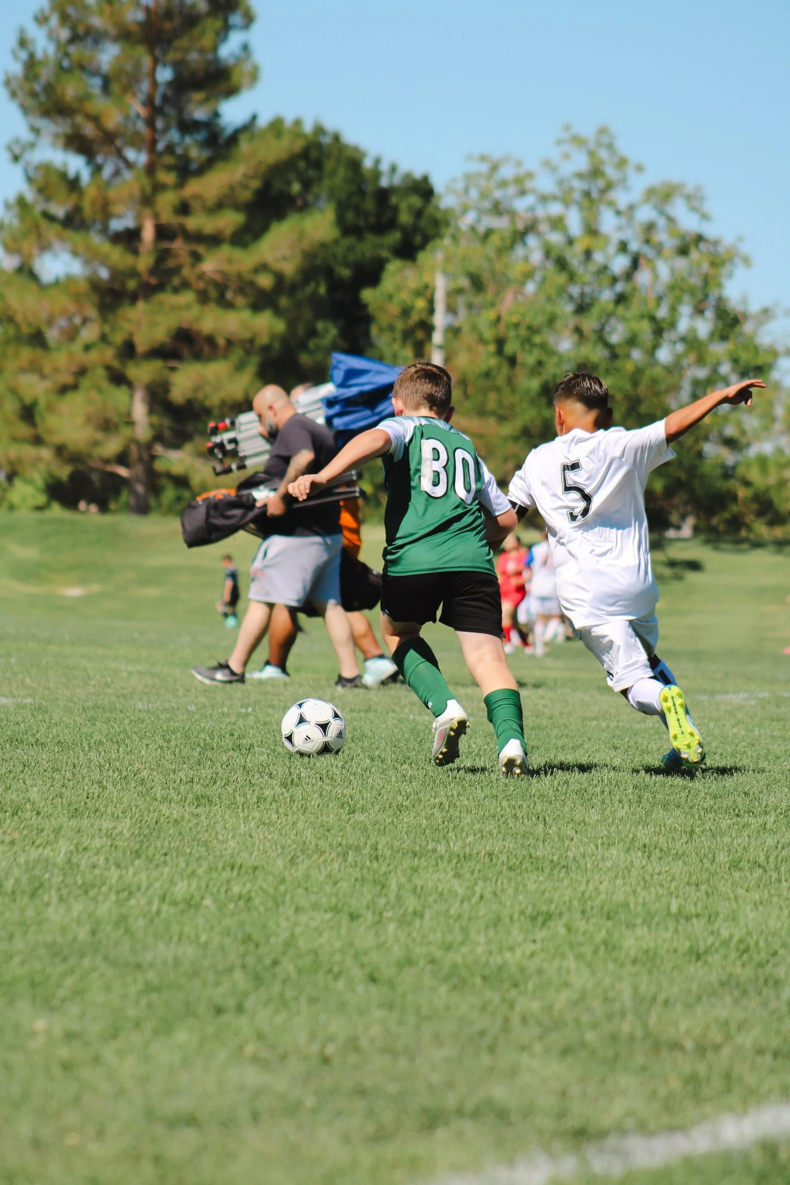 Two young soccer players running after the ball on a grassy field with trees in the background.