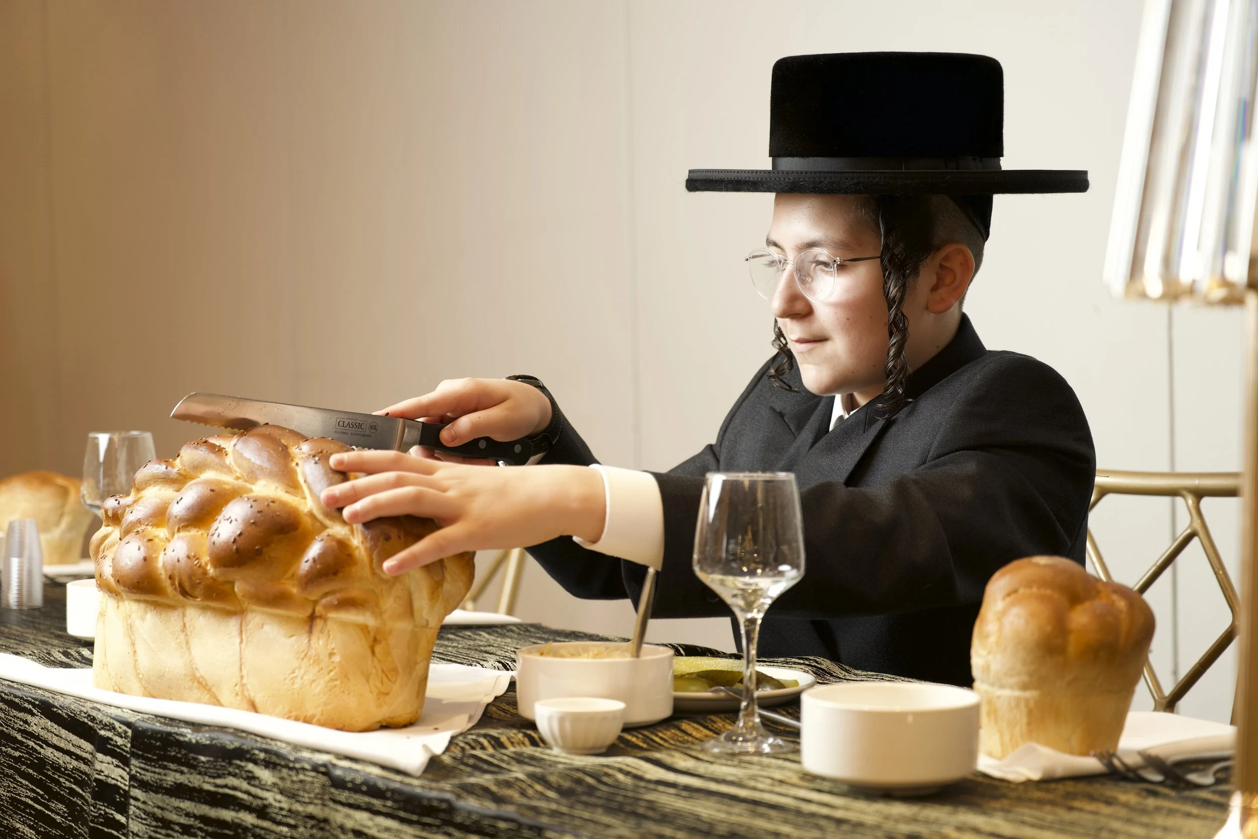 Person in a dark suit, glasses, and a black hat with a wide brim, using a knife to cut a large loaf of bread at a table set with wine glasses, small bowls, and other bread loaves in a well-lit room