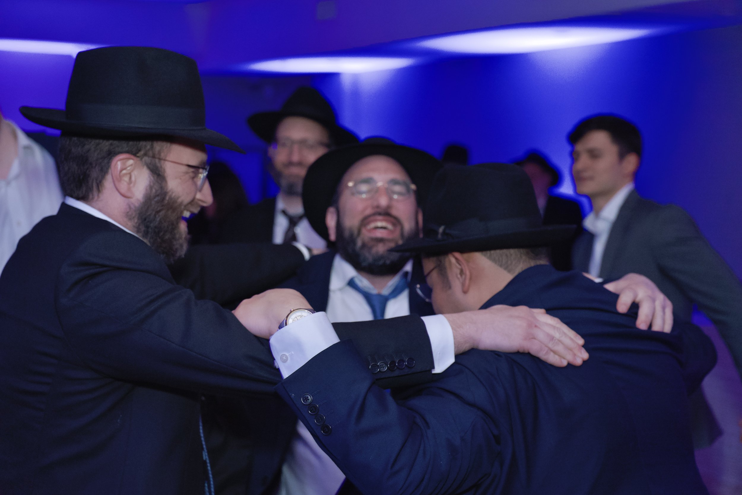 Group of men in suits and hats hugging and celebrating at an indoor event with purple lighting.