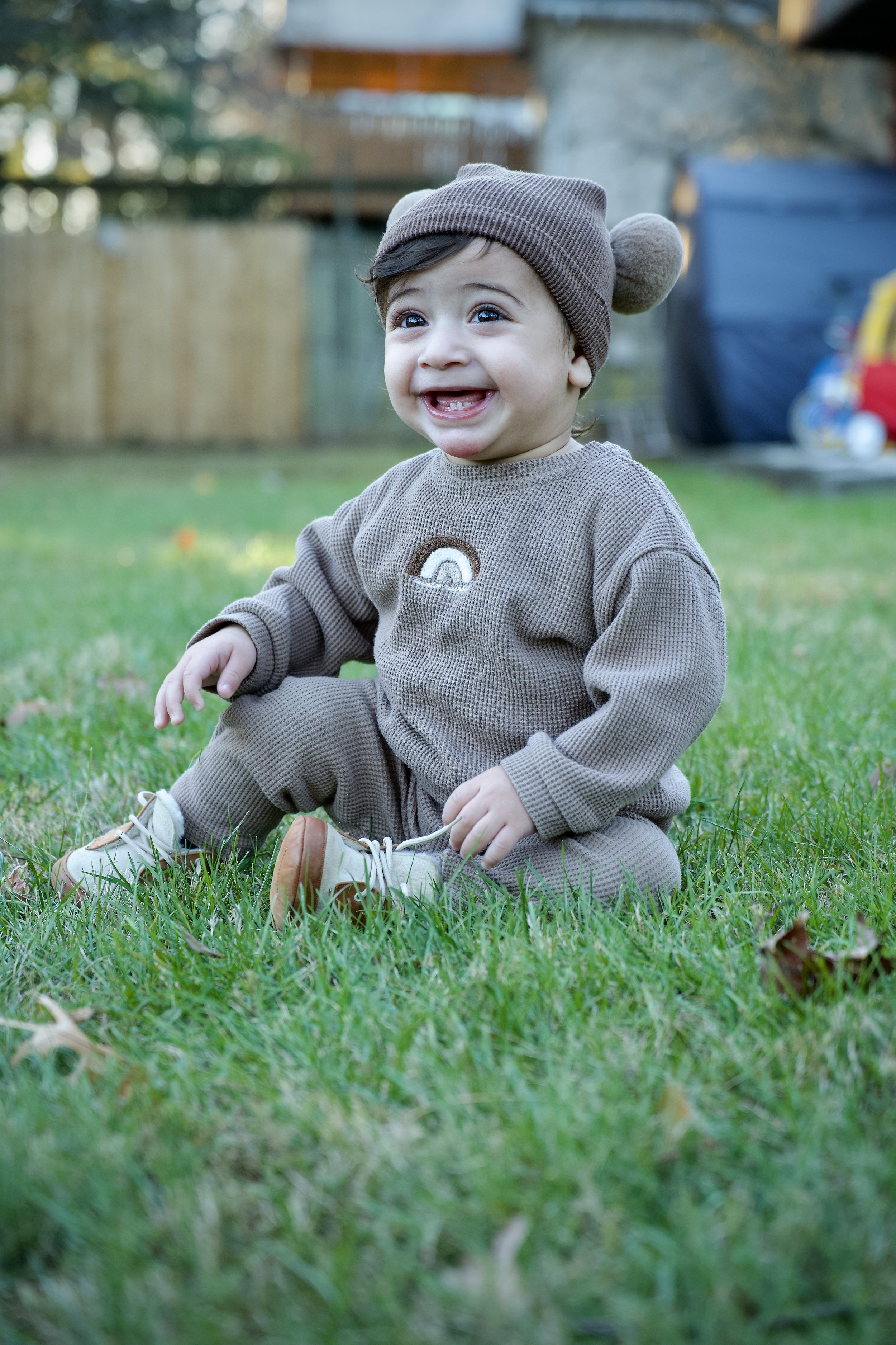 A young child sitting on grass in a backyard, smiling and wearing a brown hat with pom-poms, a matching sweater with a rainbow patch, and beige pants and shoes.
