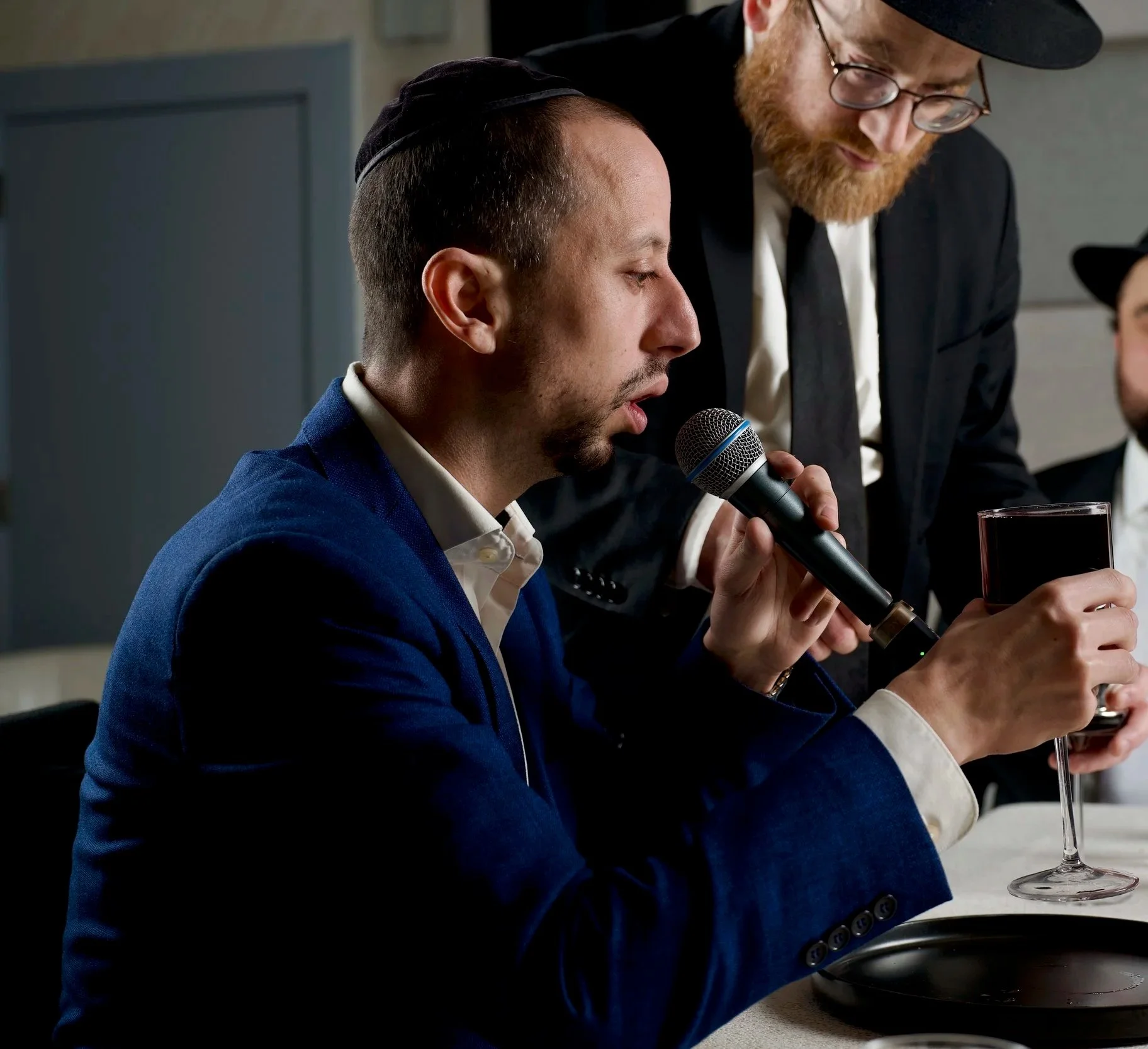 A man in a blue suit is sitting at a table, speaking into a microphone. Beside him, another man with a red beard, glasses, and a black hat is leaning over, holding a glass of red wine, and looking at the man speaking.