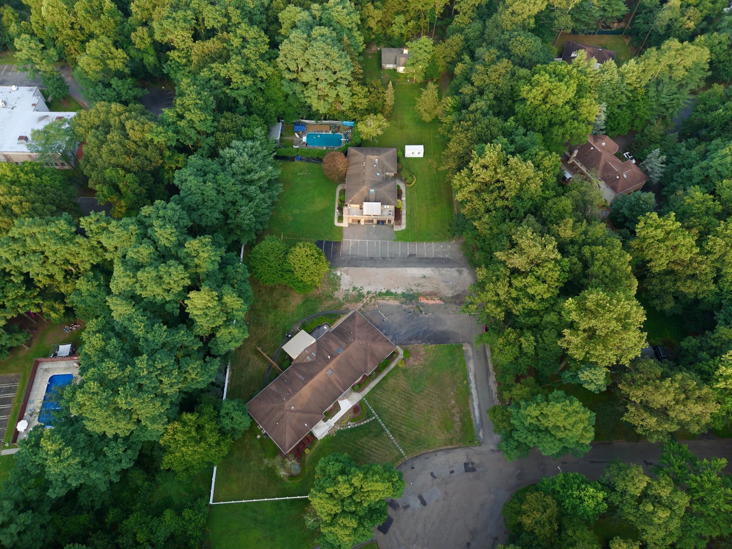 An aerial view of residential houses surrounded by tall green trees, with a backyard featuring a swimming pool and a lawn, and a parking area at the bottom of the image.