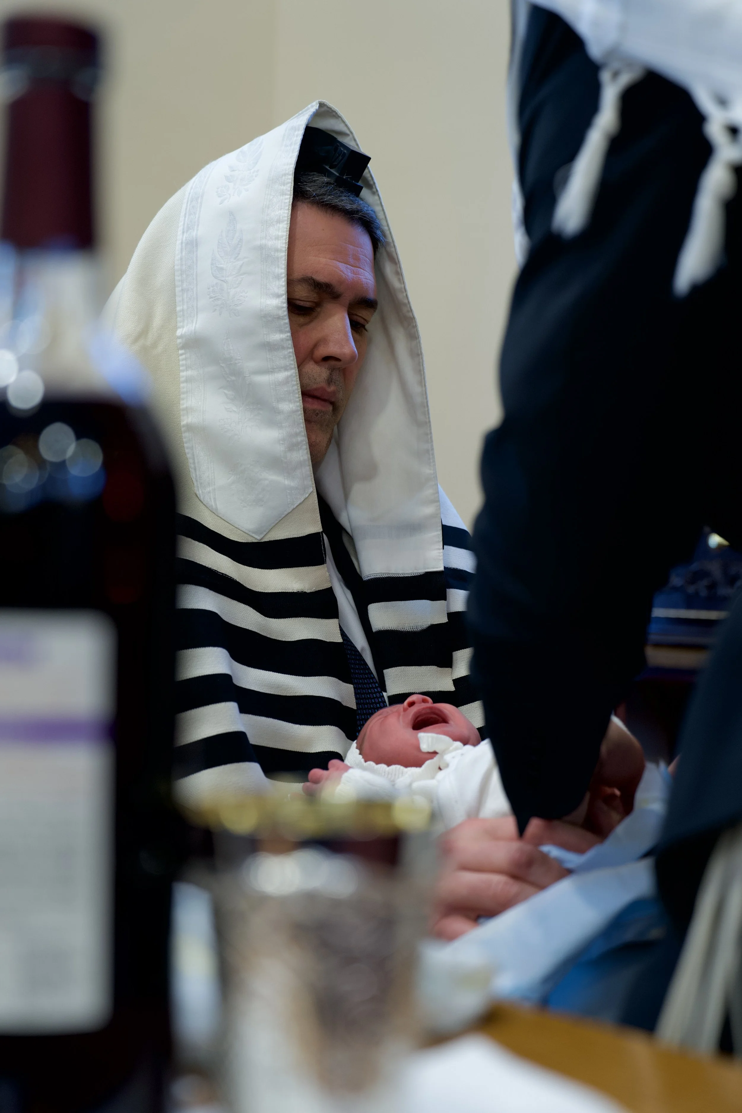 A man wearing a white garment and a prayer shawl cradles a crying baby in a clinical setting, with medical staff nearby.
