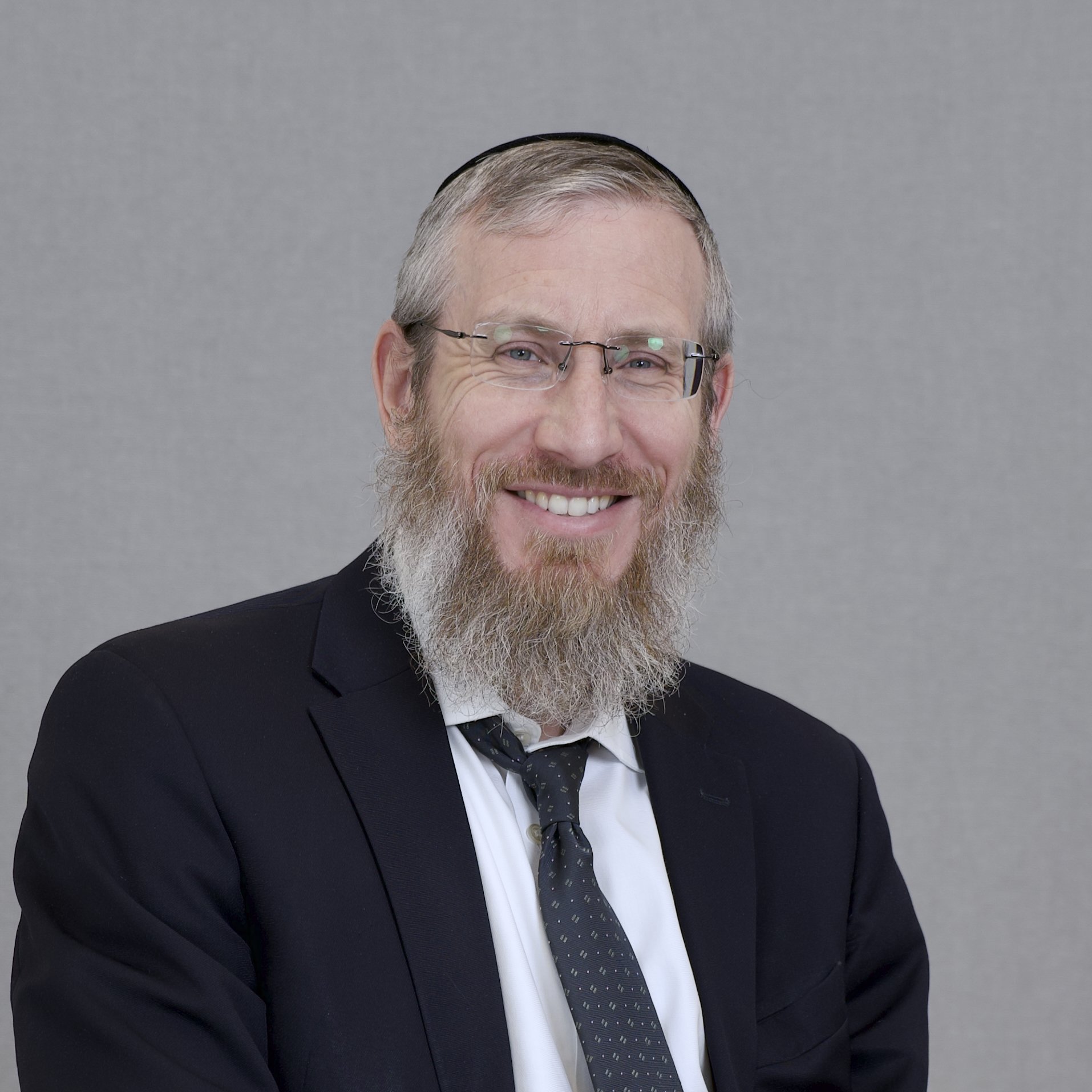 Smiling man with glasses, gray hair, and beard wearing a black suit and patterned tie, posing in front of a plain gray background.