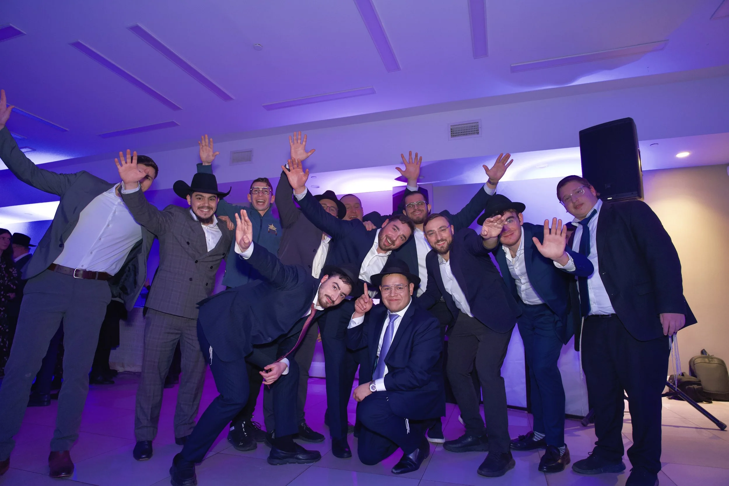 Group of people celebrating at an indoor event, some raising their hands and smiling, wearing formal and semi-formal attire with some in suits and hats, purple lighting highlights the festive scene.