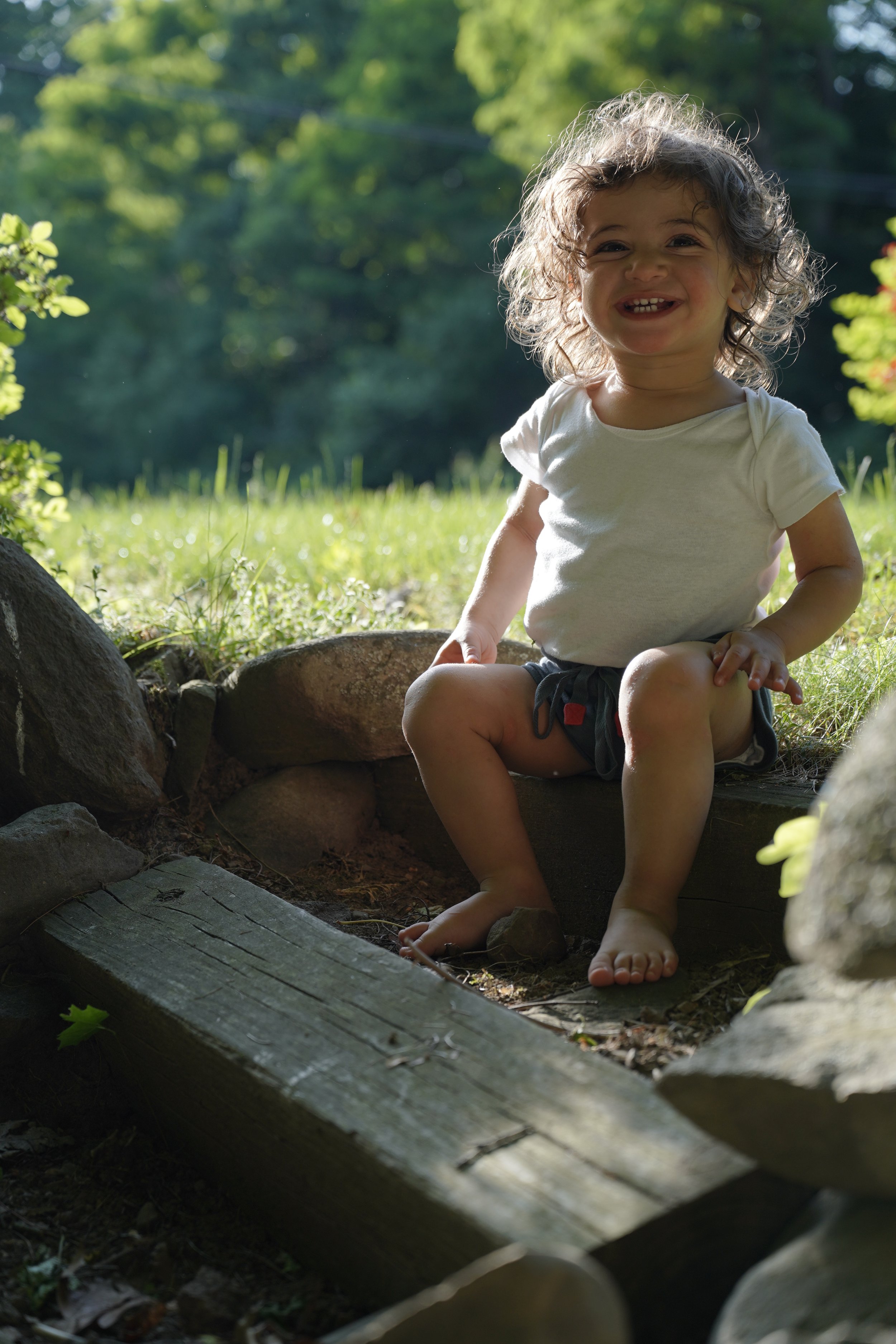 A young girl with curly brown hair, smiling and showing her teeth, sitting outdoors on a large rock with greenery and trees in the background.