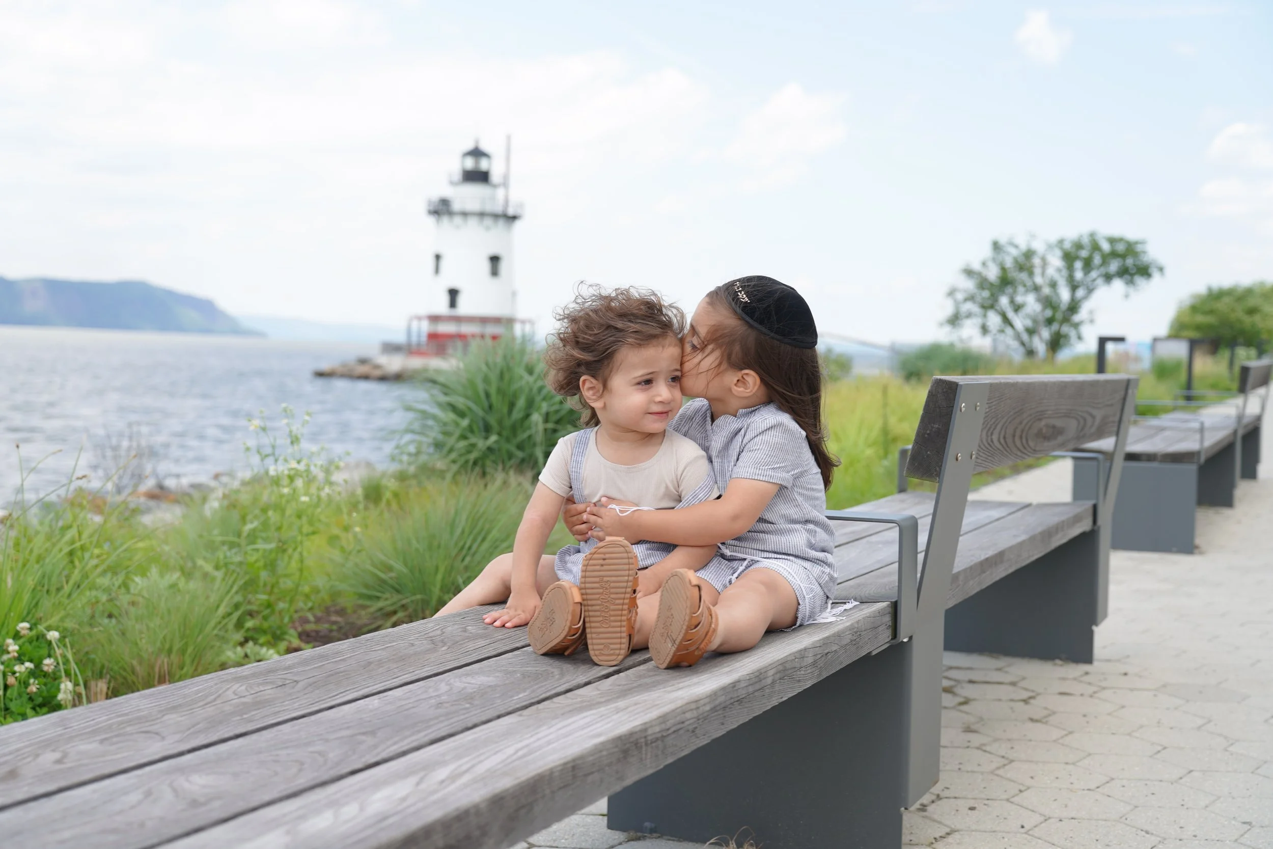 A woman and a young boy sitting on a wooden bench outdoors near water, with a lighthouse in the background. The woman is kissing the boy on the cheek, and they both appear happy. Green grass and trees are also visible.