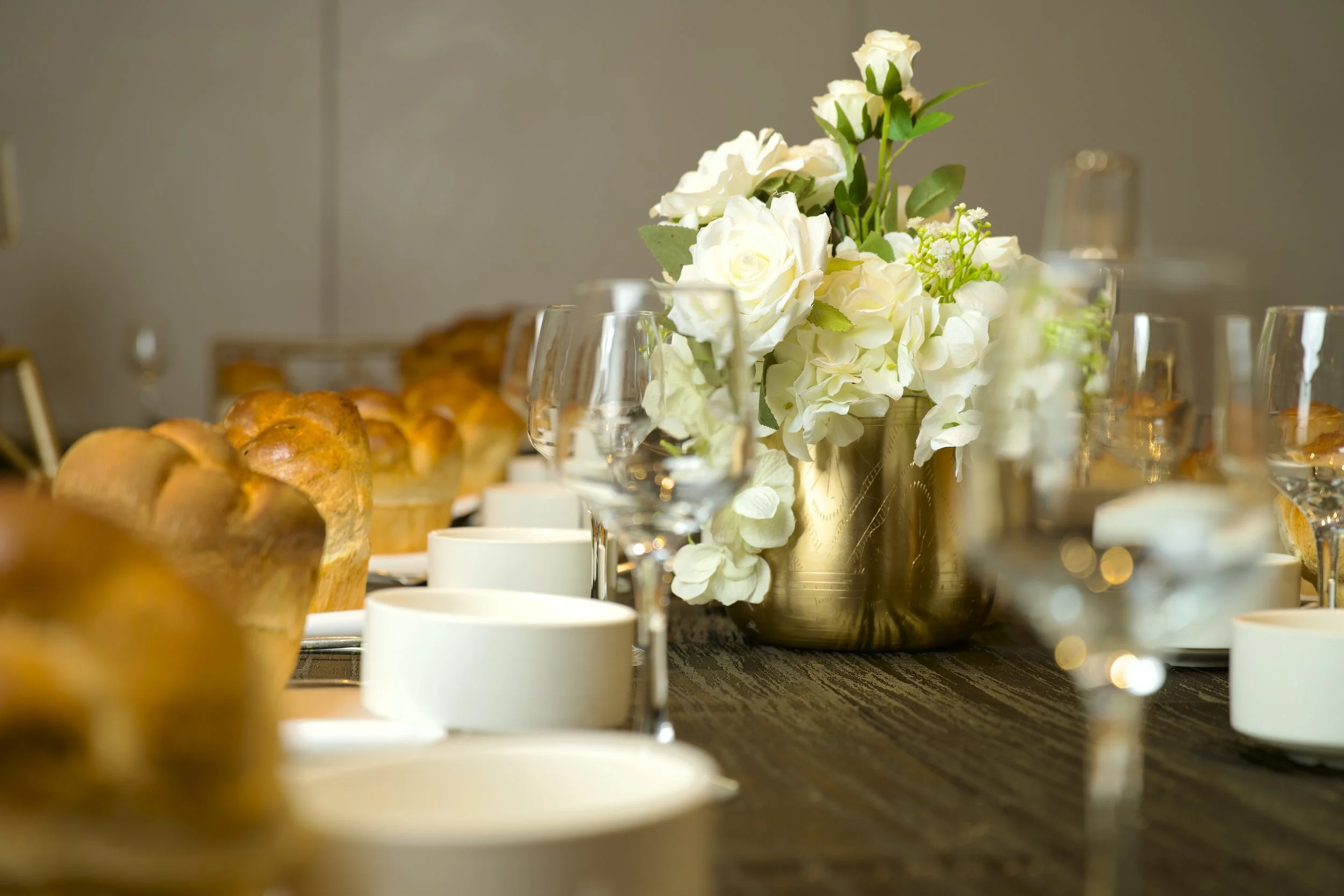 A table decorated with a white floral centerpiece, white cups, glasses, and baked goods in the background.