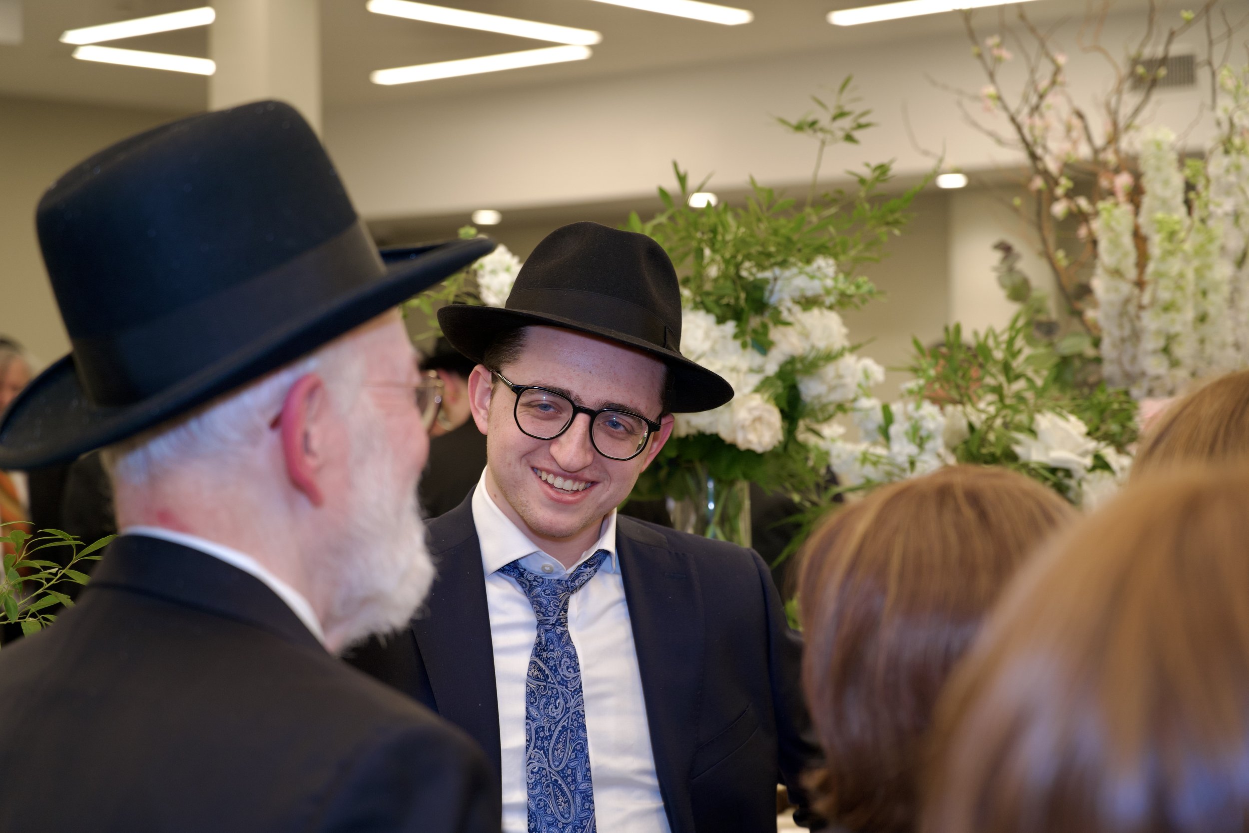 People gathered at an event, wearing formal attire, with a background of floral arrangements and modern lighting.