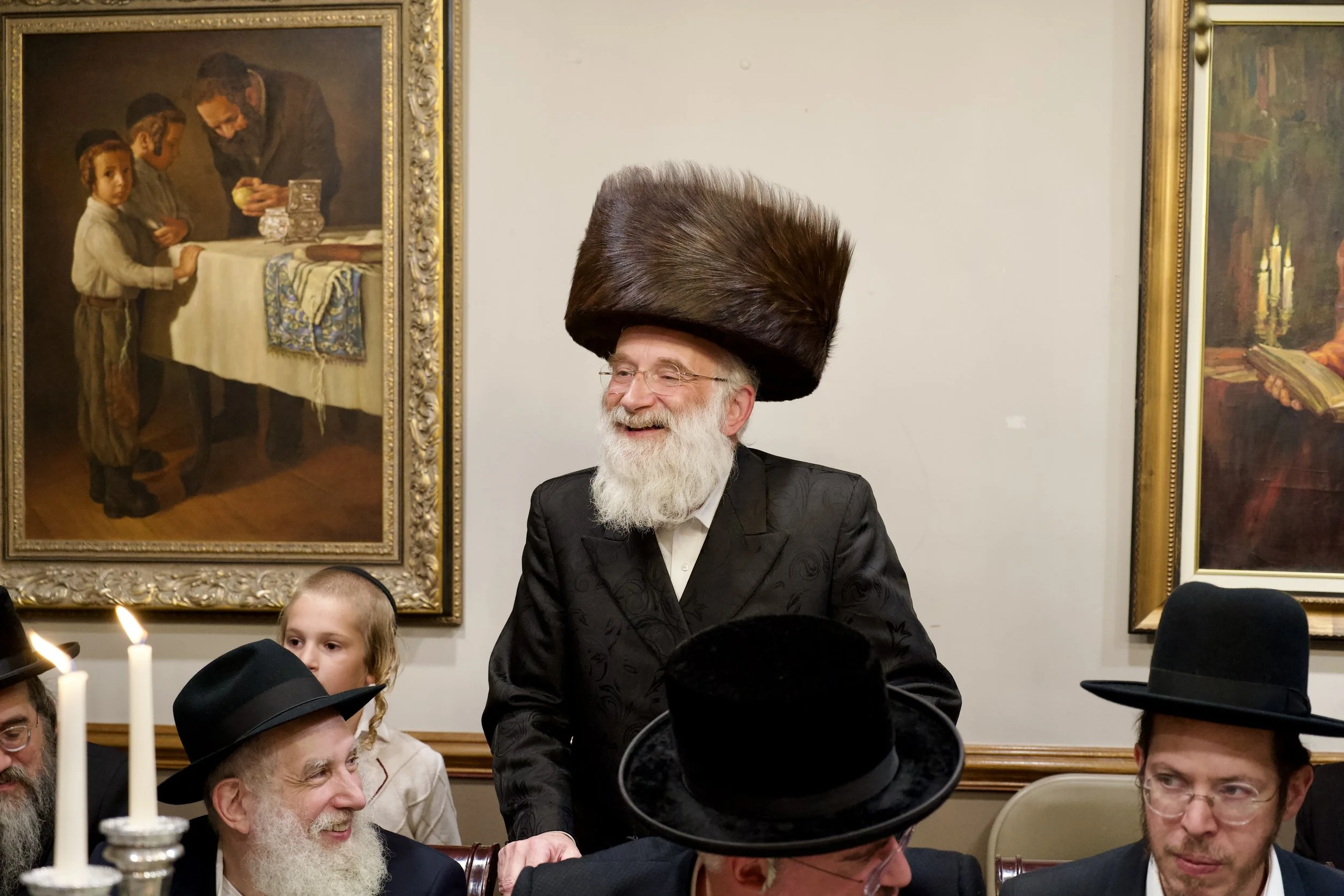 A cheerful man with a white beard and glasses, dressed in a black suit, wearing a large fur hat, standing among others at a gathering, with paintings on the wall behind him.