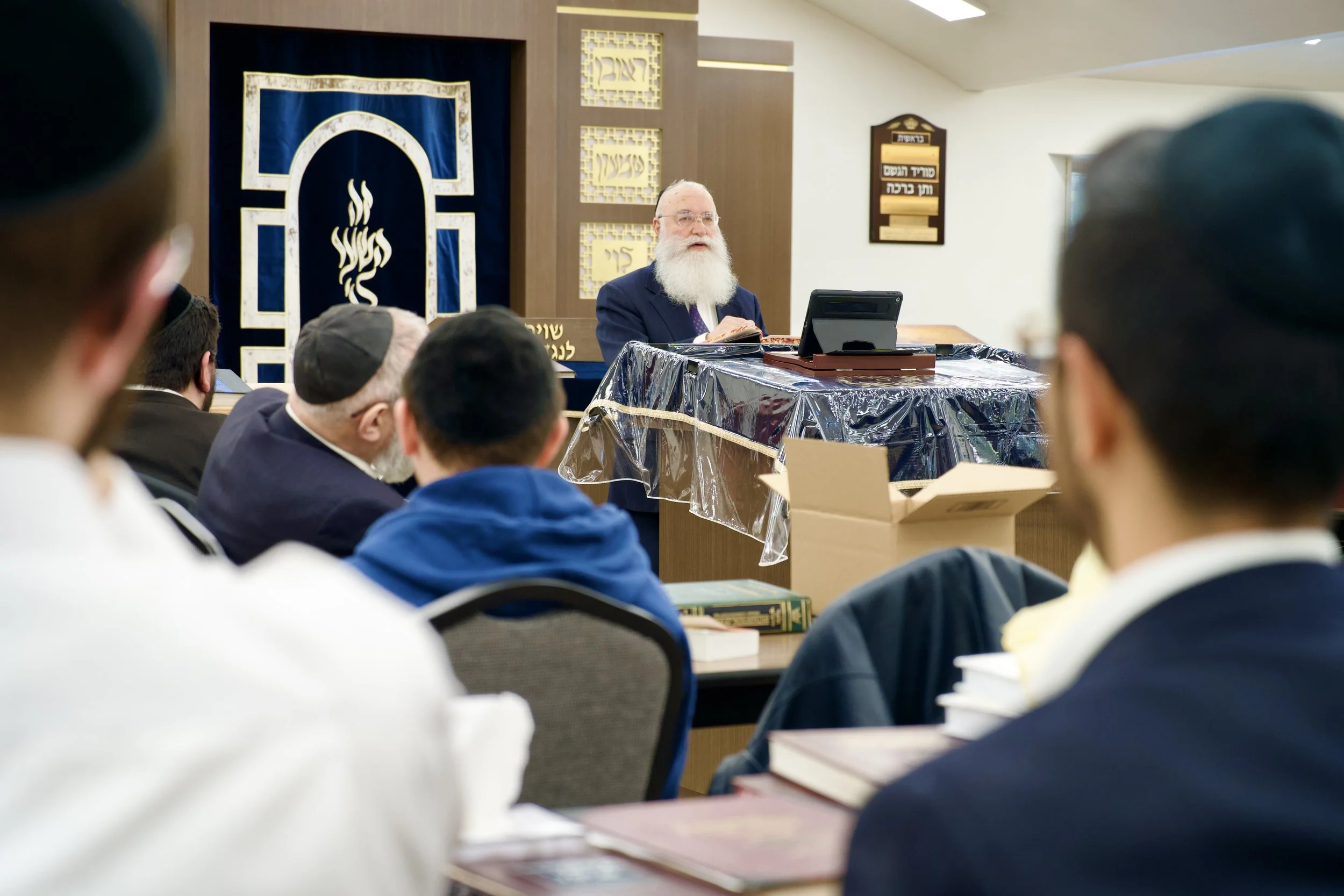 A Jewish rabbi with a long white beard and glasses, dressed in a blue suit, is speaking or teaching at a gathering. He is seated at a table with a book and a digital device, in a room decorated with Hebrew signs and symbols, including a blue curtain 