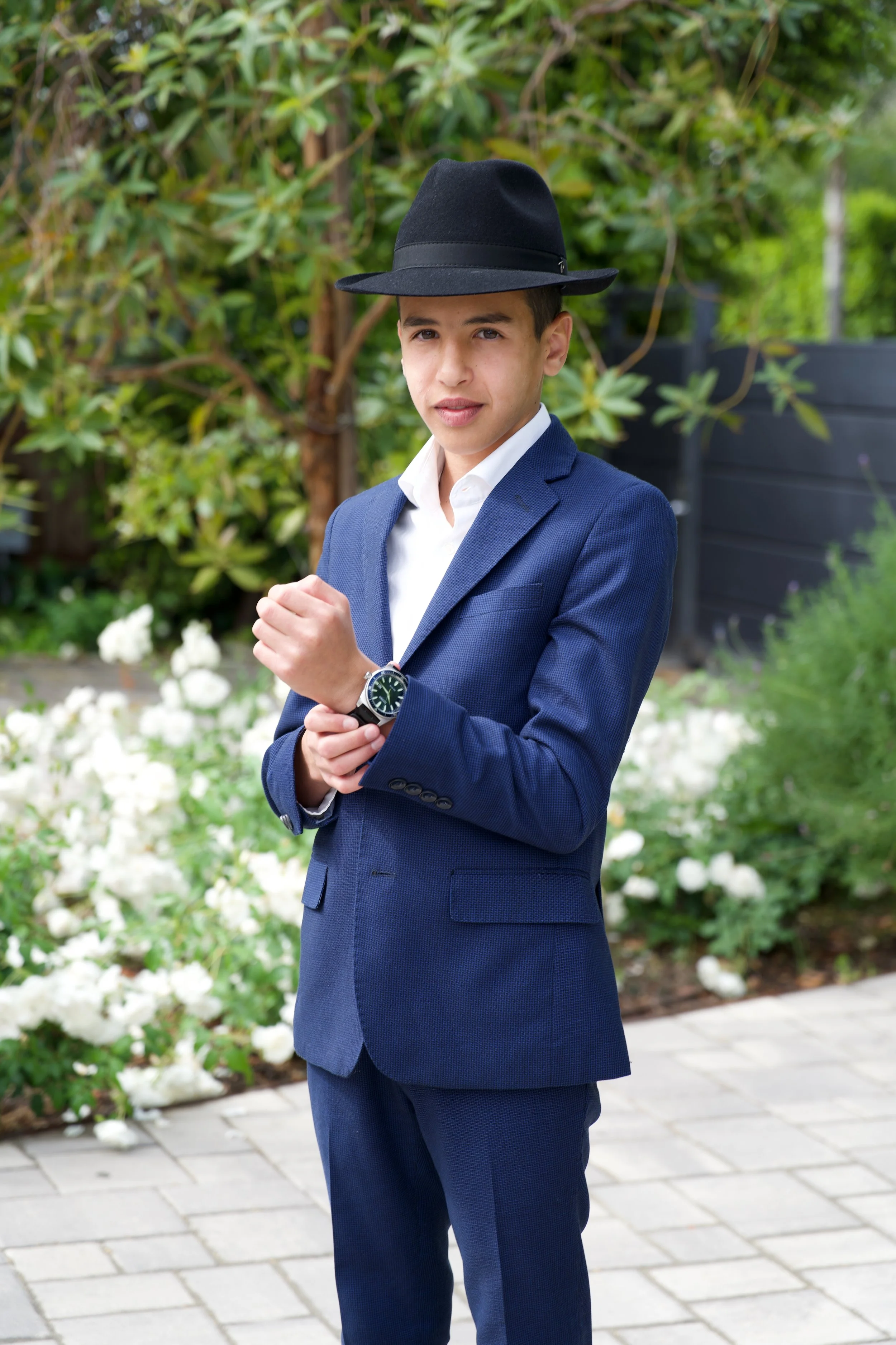 A young man wearing a navy blue suit, white shirt, black watch, and black fedora hat, standing outdoors on a paved walkway with green bushes and white flowers in the background.