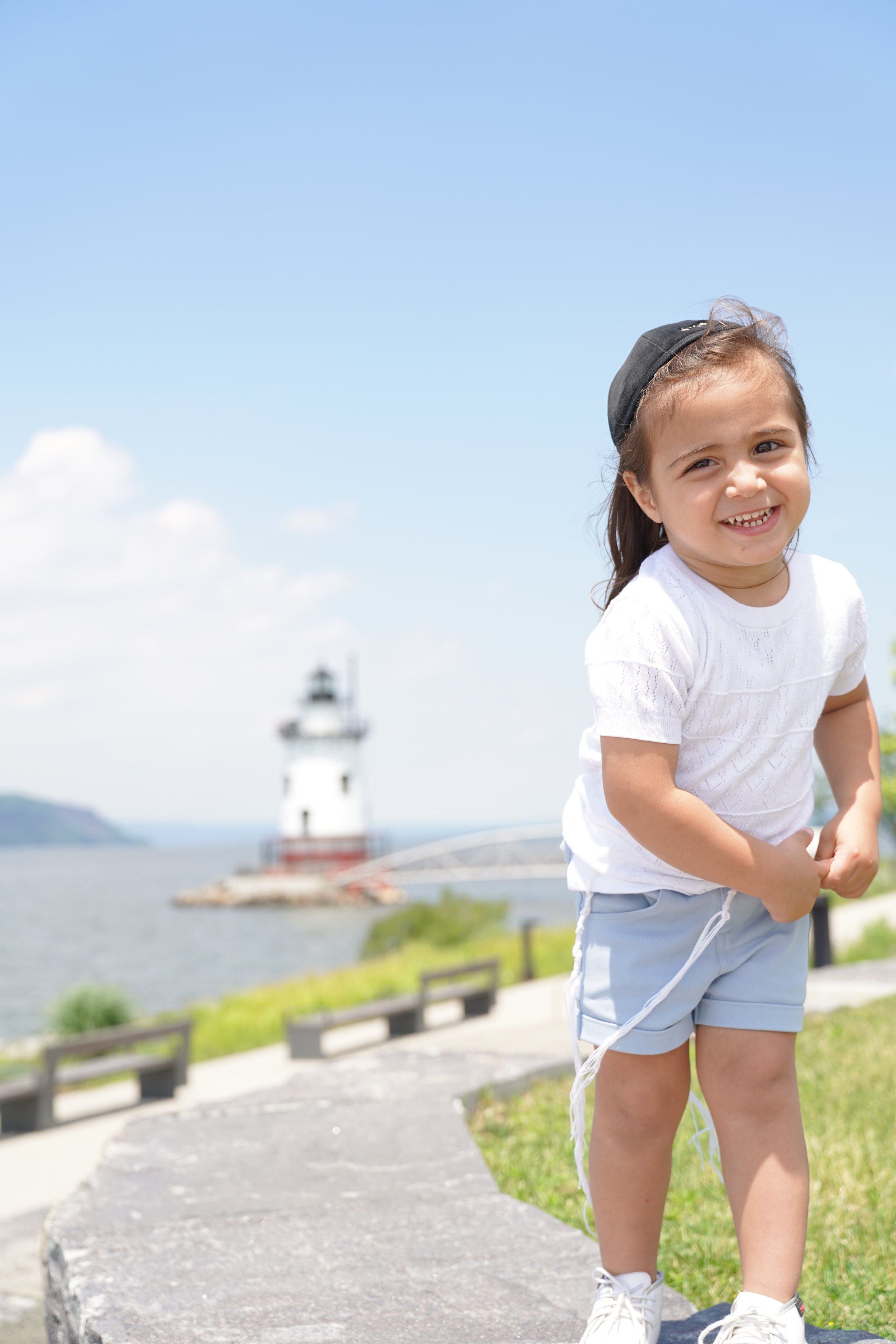 A smiling young girl in a white T-shirt, light blue shorts, and white sneakers, standing outdoors near a lighthouse with a blue sky and scattered clouds in the background.