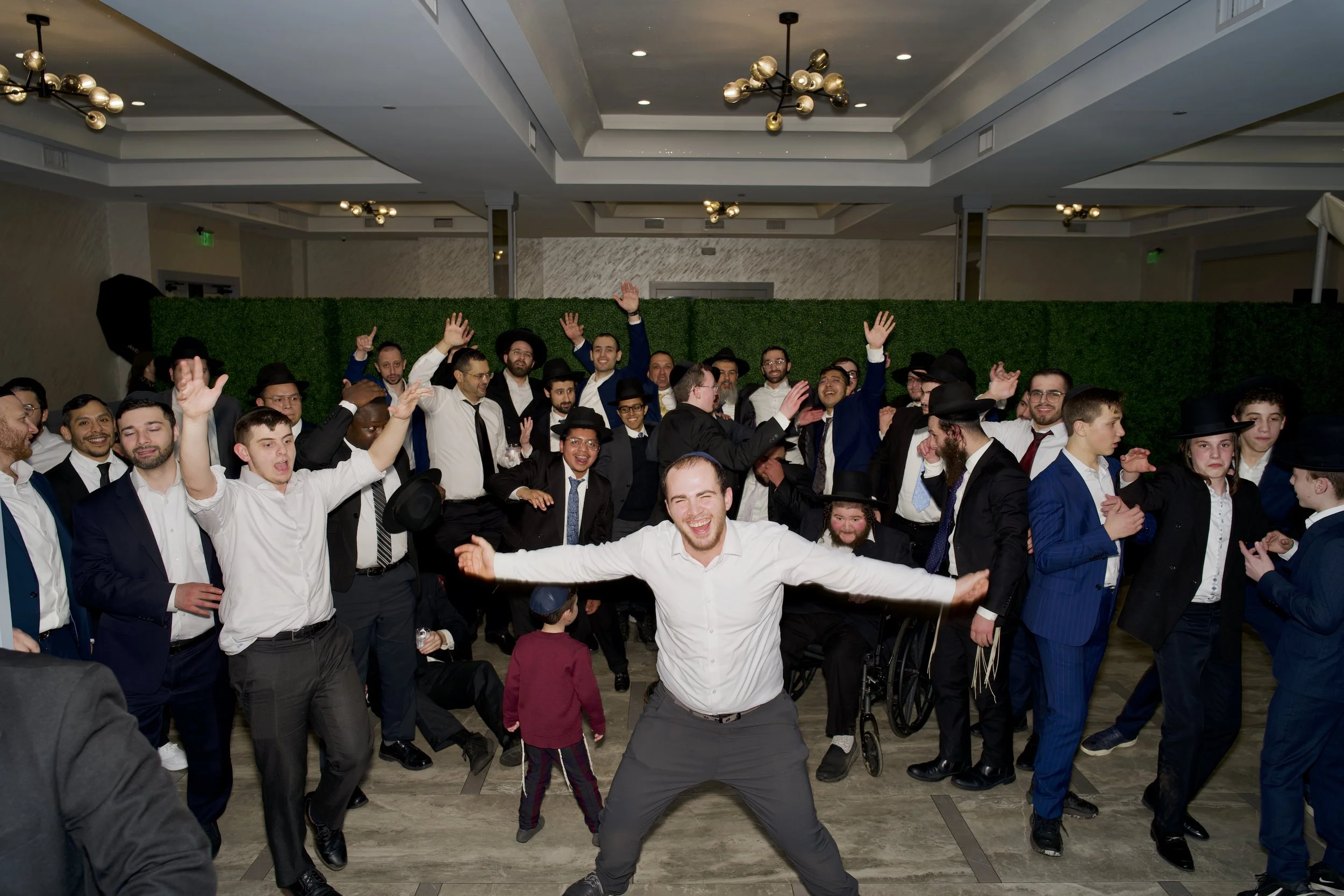 A group of men and boys dressed in formal attire, celebrating and dancing at an indoor event, with some raising their hands and smiling.