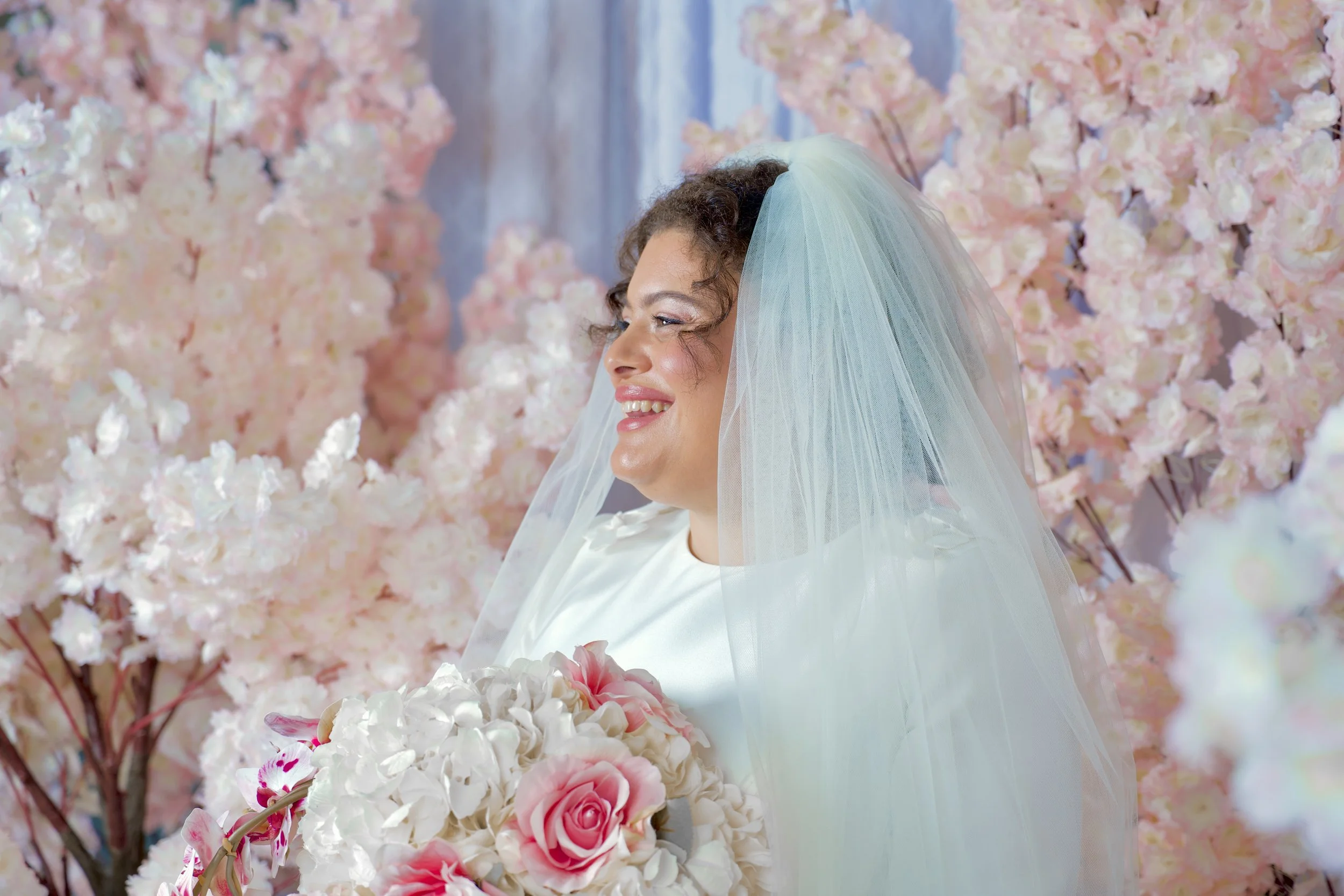 A person wearing a light blue veil and white attire, smiling, with pink and white flowers and pink cherry blossom trees in the background.
