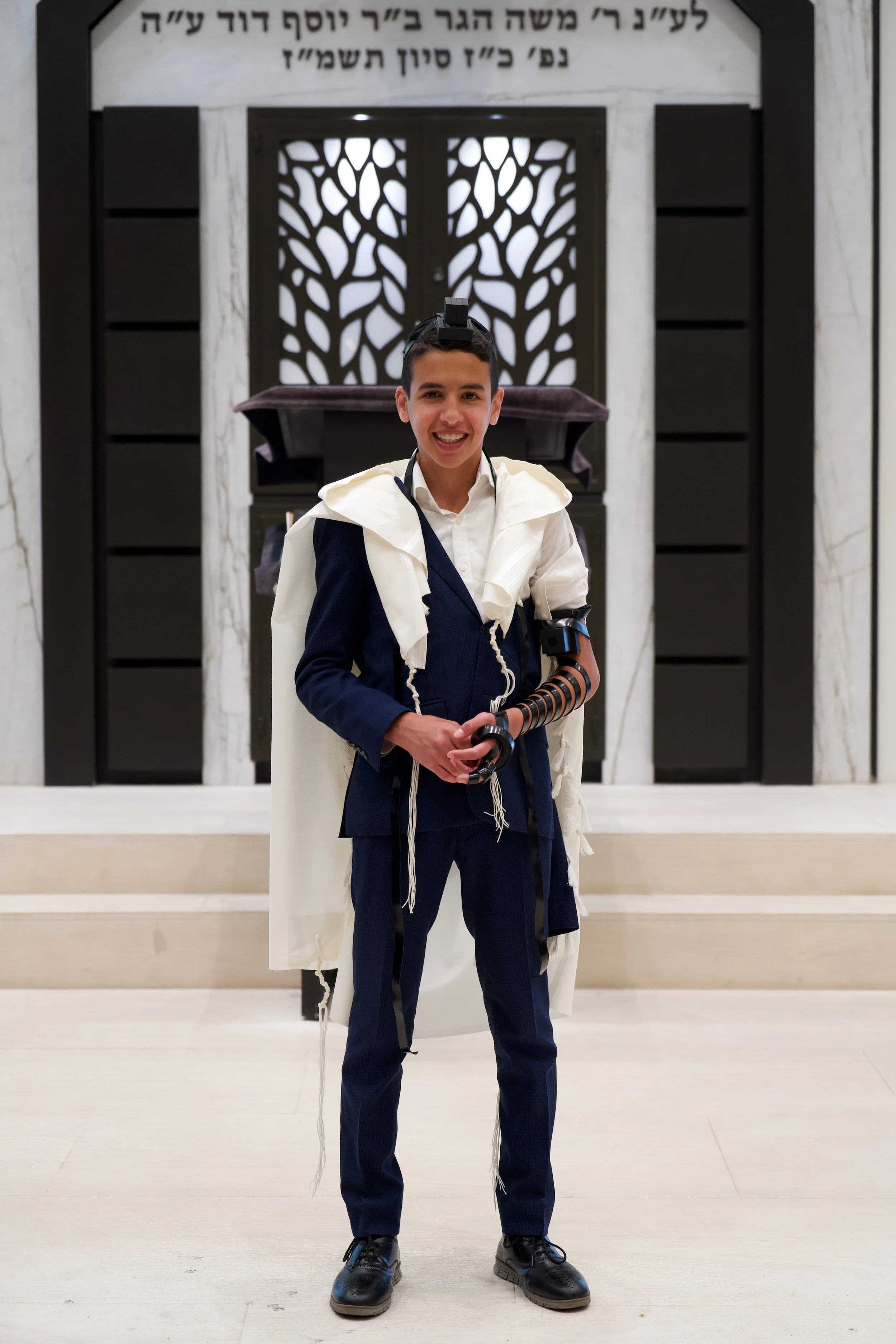 A young boy in formal attire wearing a white ceremonial prayer shawl and a black kippah with a device on his head, standing inside a synagogue. The background features an ornate wooden ark with Hebrew inscriptions.