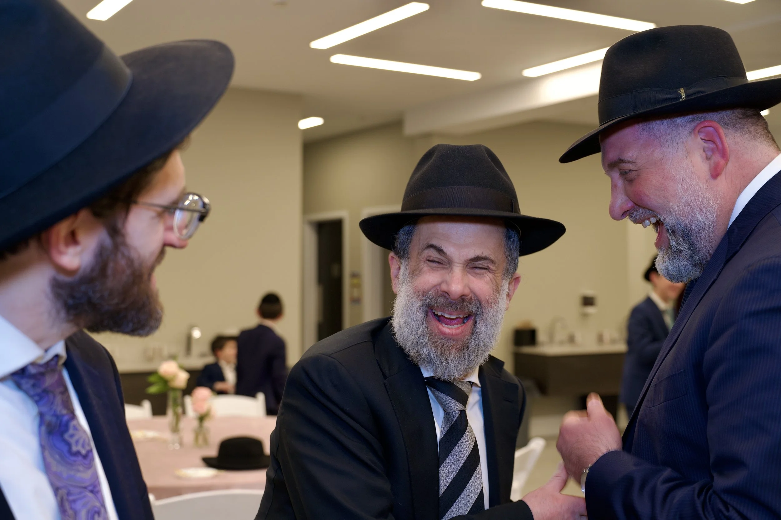 Three men in formal attire, including black hats, are engaged in conversation and laughing at a social event in a well-lit room with decorated tables and flowers.