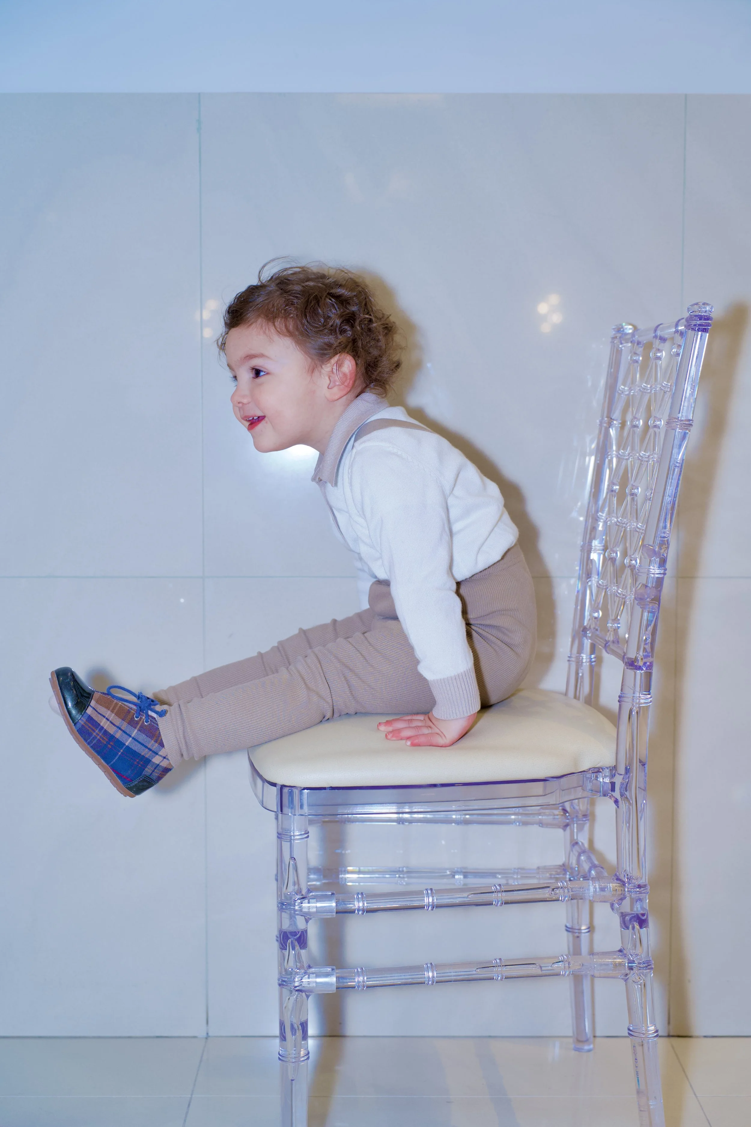 A young boy with curly hair is sitting sideways on a transparent chair, leaning back against the wall, with his legs extended and hands resting on the chair. He is wearing a light-colored sweater, beige pants, and plaid shoes, and is smiling while lo