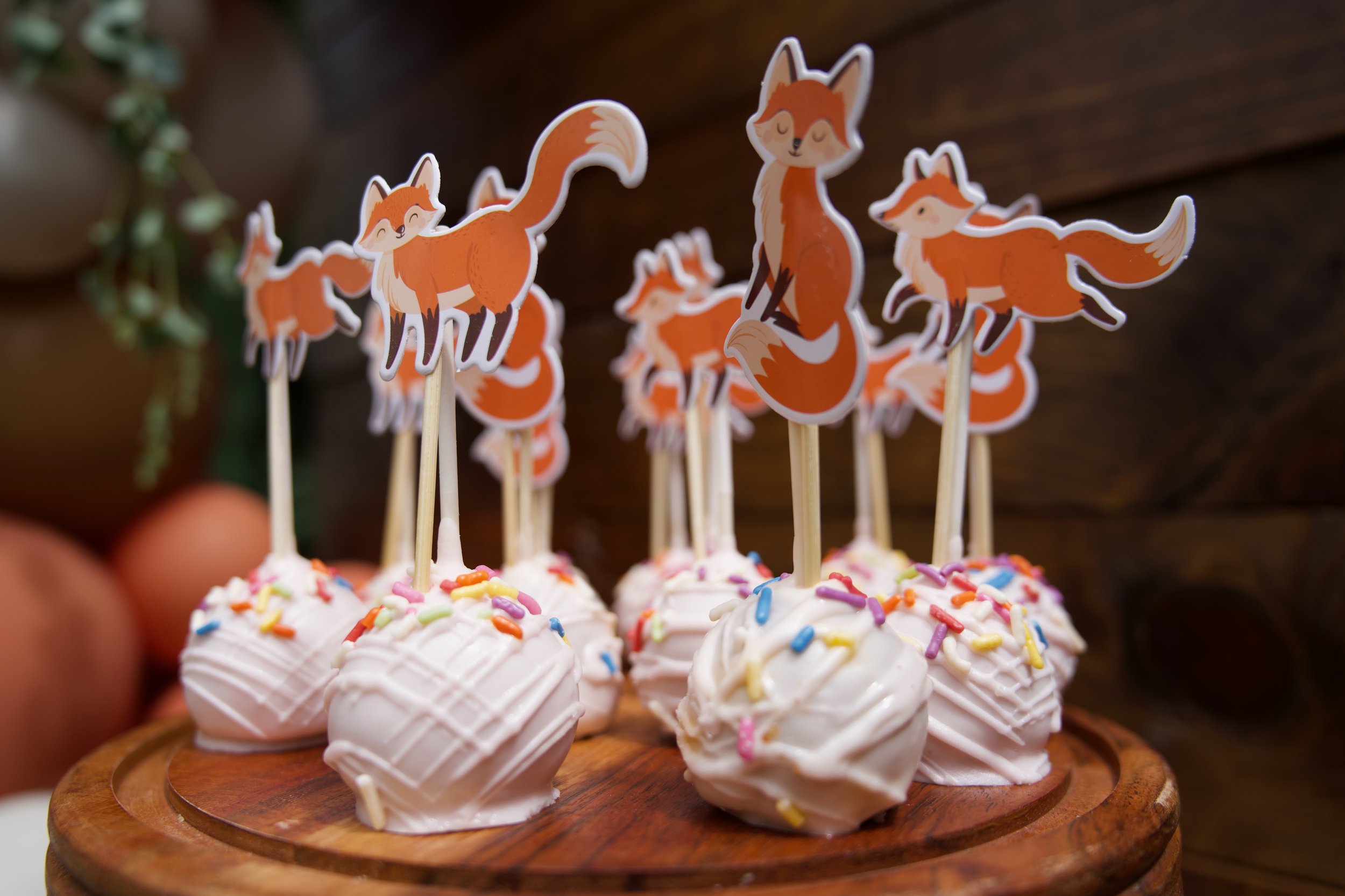 Cupcakes with white frosting and colorful sprinkles topped with fox-shaped toppers on sticks, arranged on a wooden serving tray.