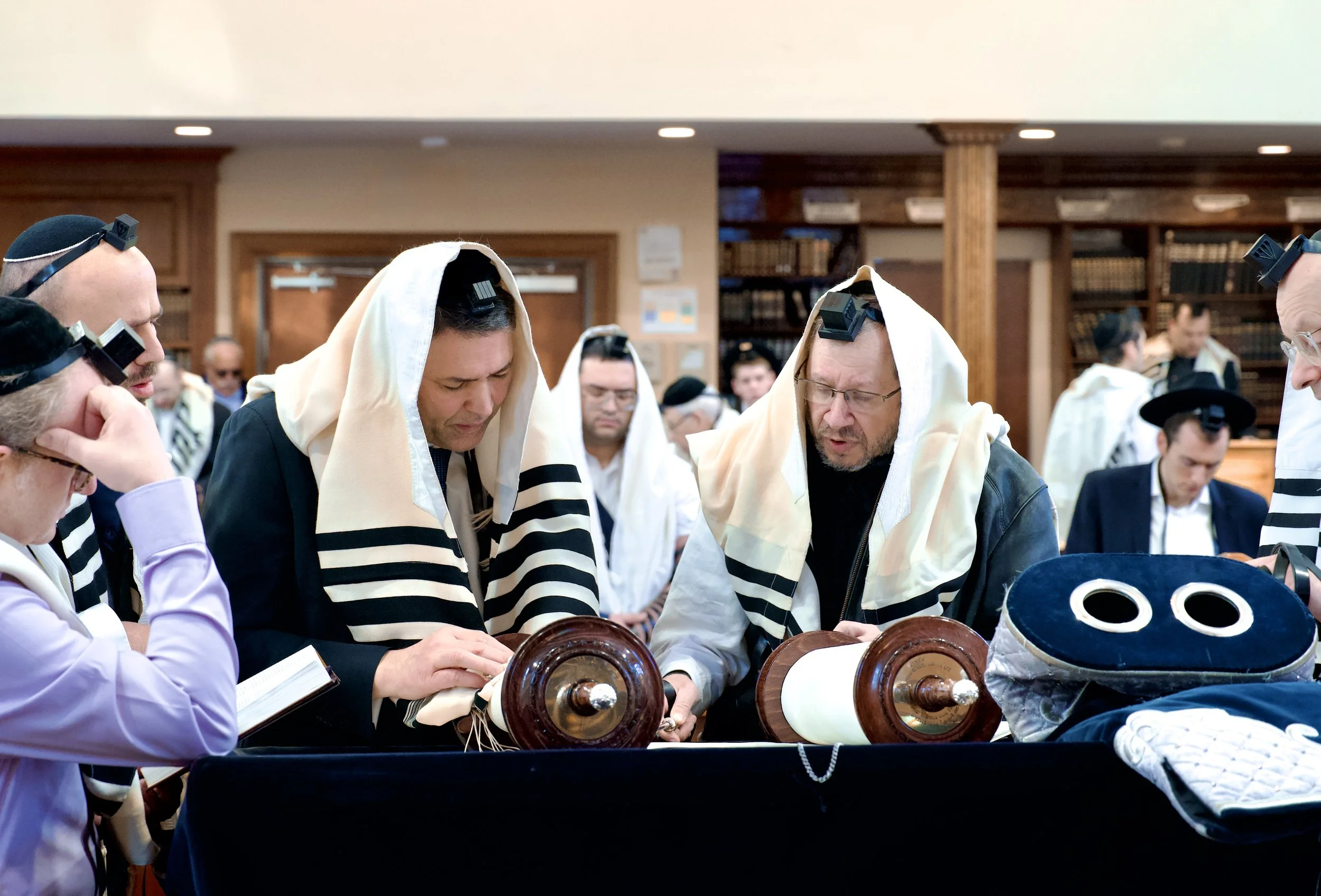 Group of men wearing traditional Jewish prayer shawls and head coverings, gathered around a table, reading from religious texts in a synagogue or study hall.