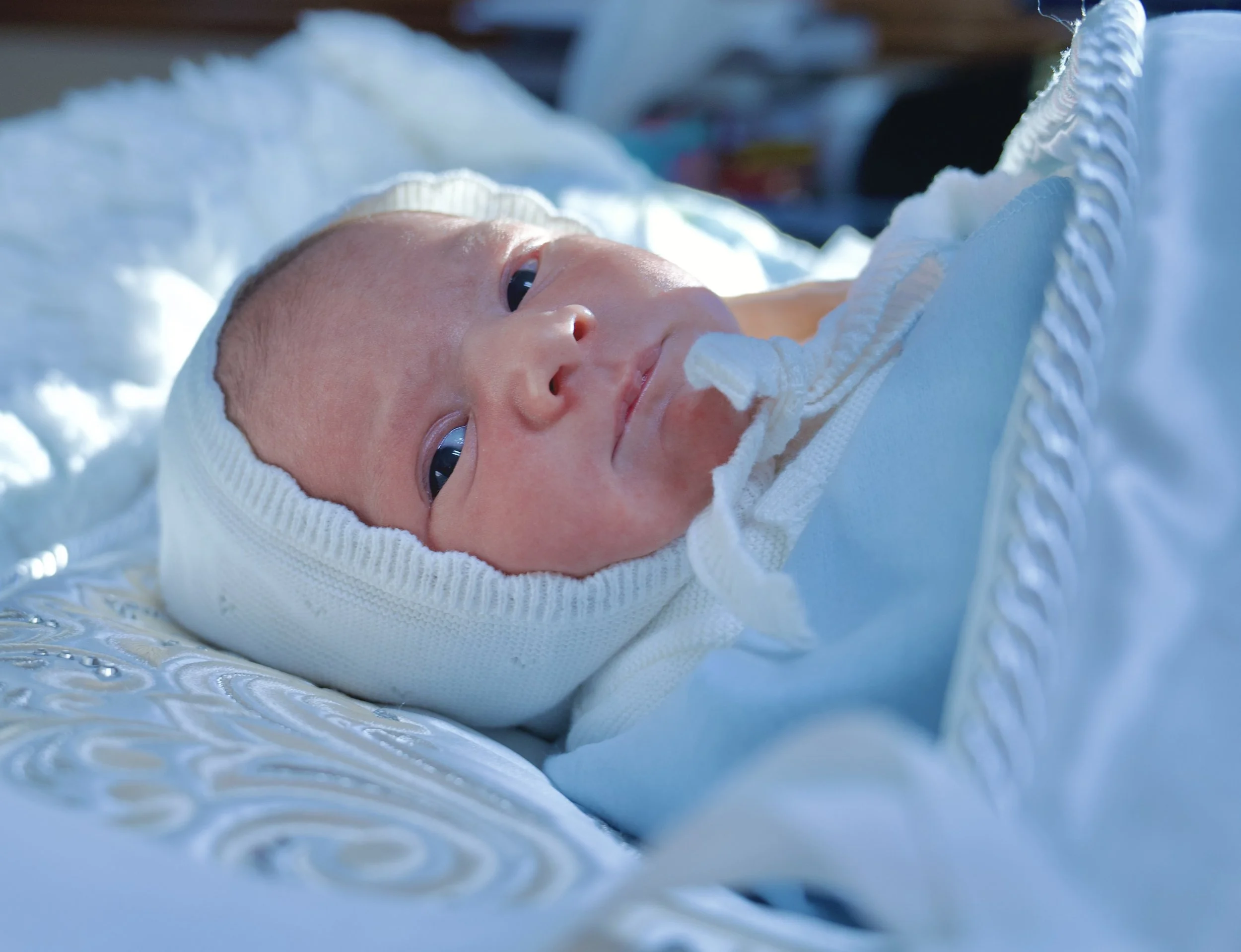 Close-up of a newborn baby lying on a hospital bassinet covered with a white blanket and wearing a white knit hat, with an expression of curiosity.