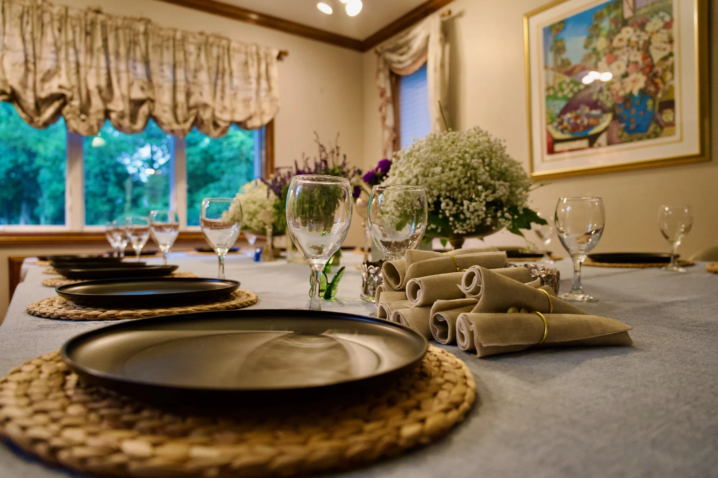 A dining room table set with placemats, wine glasses, and neatly rolled napkins. There are floral arrangements on the table, and a large framed colorful painting on the wall. The room has large windows with floral curtains, and trees are visible outs