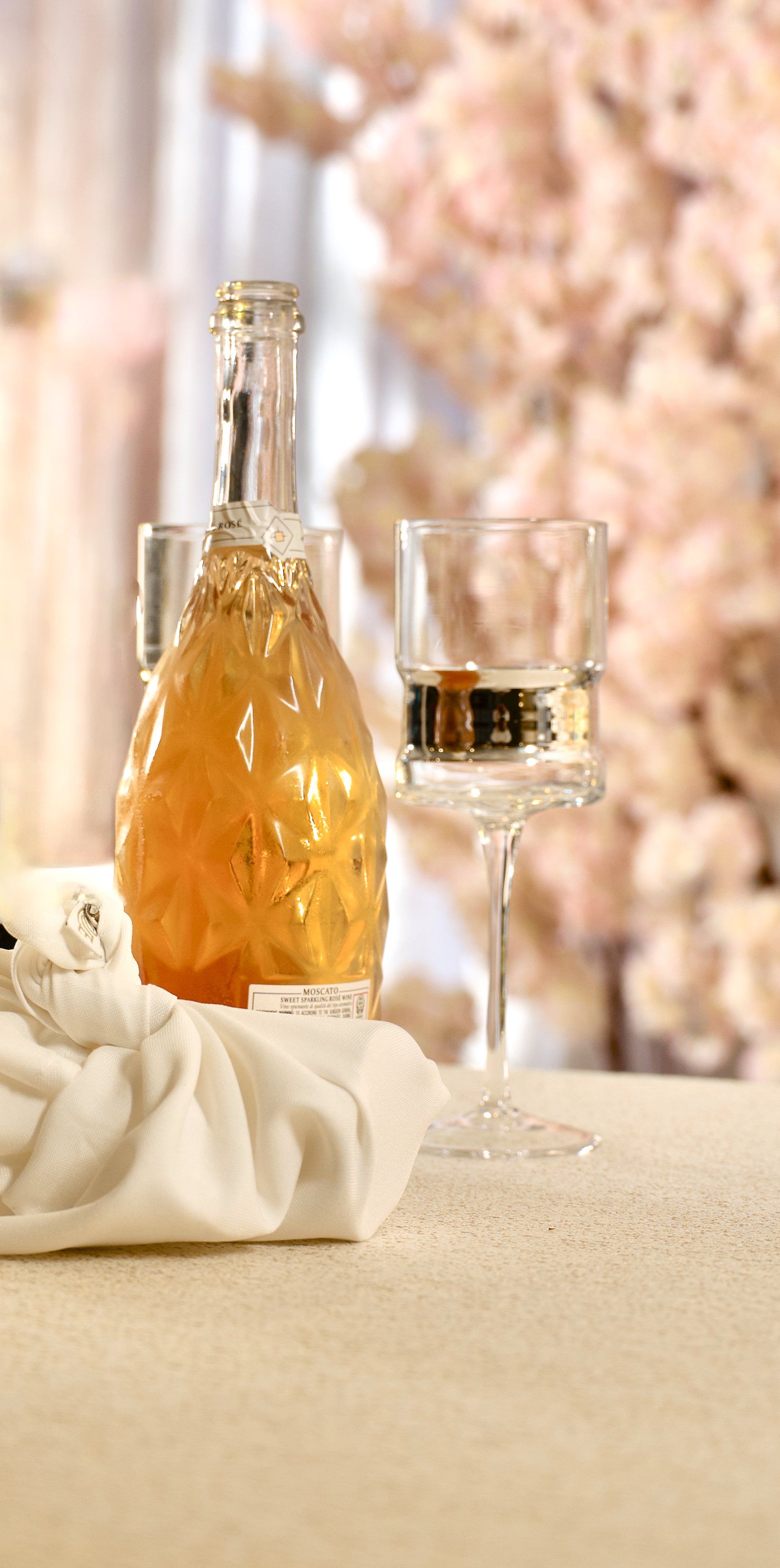 A bottle of sparkling rosé wine, a glass of water, and pink floral decorations in the background on a table.