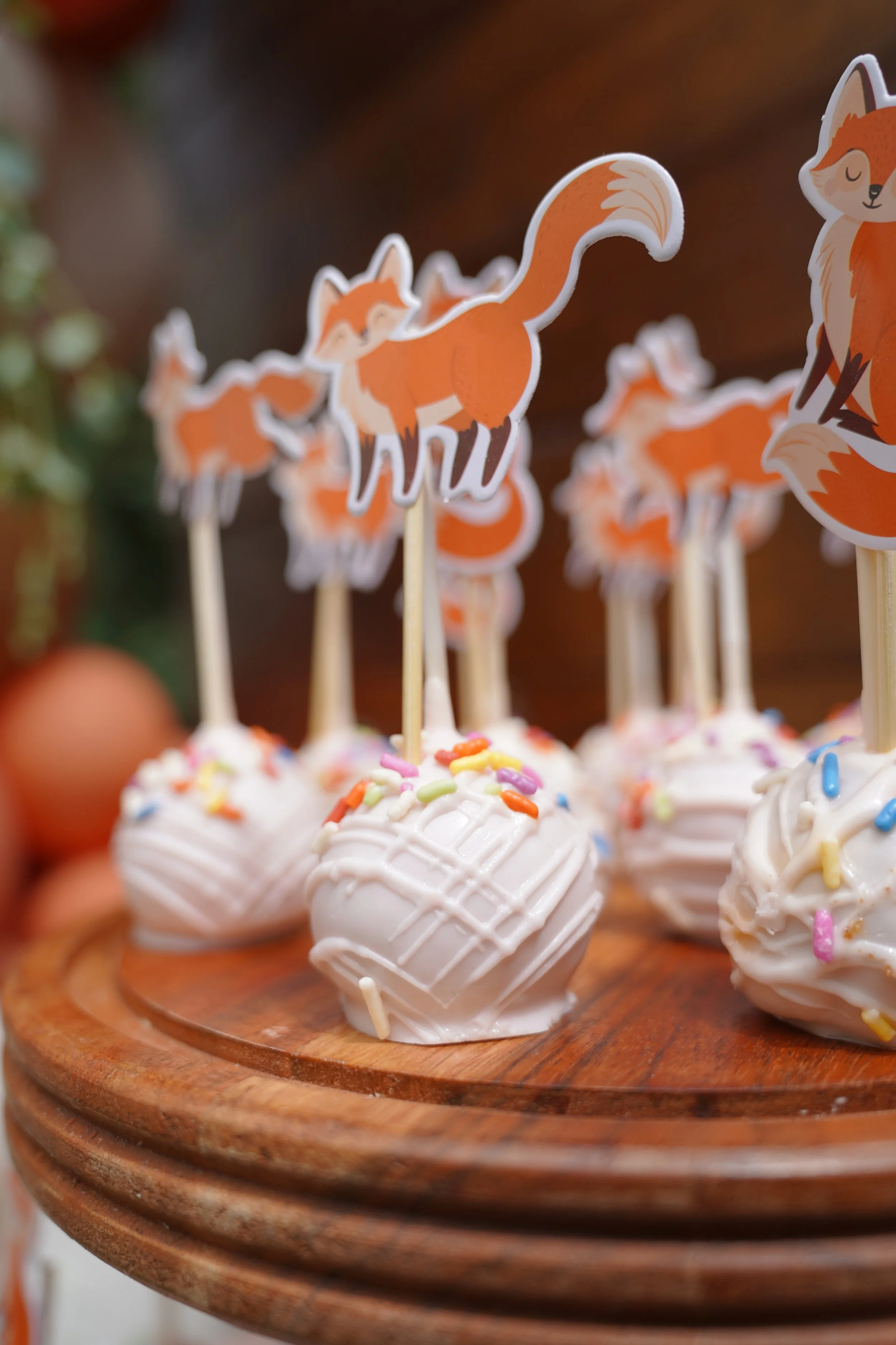 Cake pops with white frosting and colorful sprinkles on a wooden tray, decorated with fox-themed toppers.